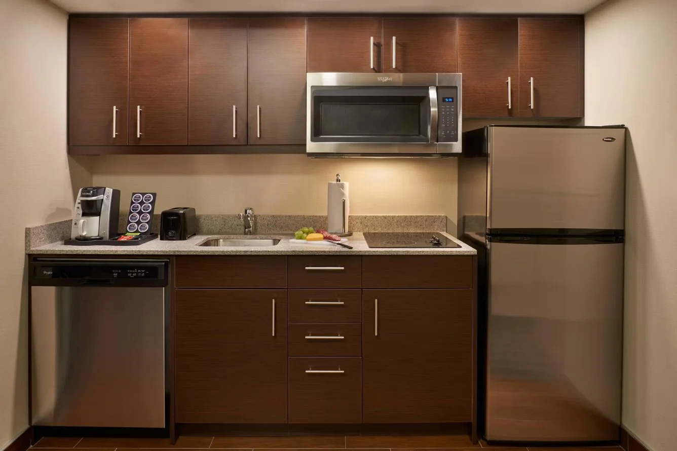 A small kitchenette with wooden cabinets, a microwave, refrigerator, dishwasher, coffee machine, toaster, sink, and a countertop with a cutting board and fruits.