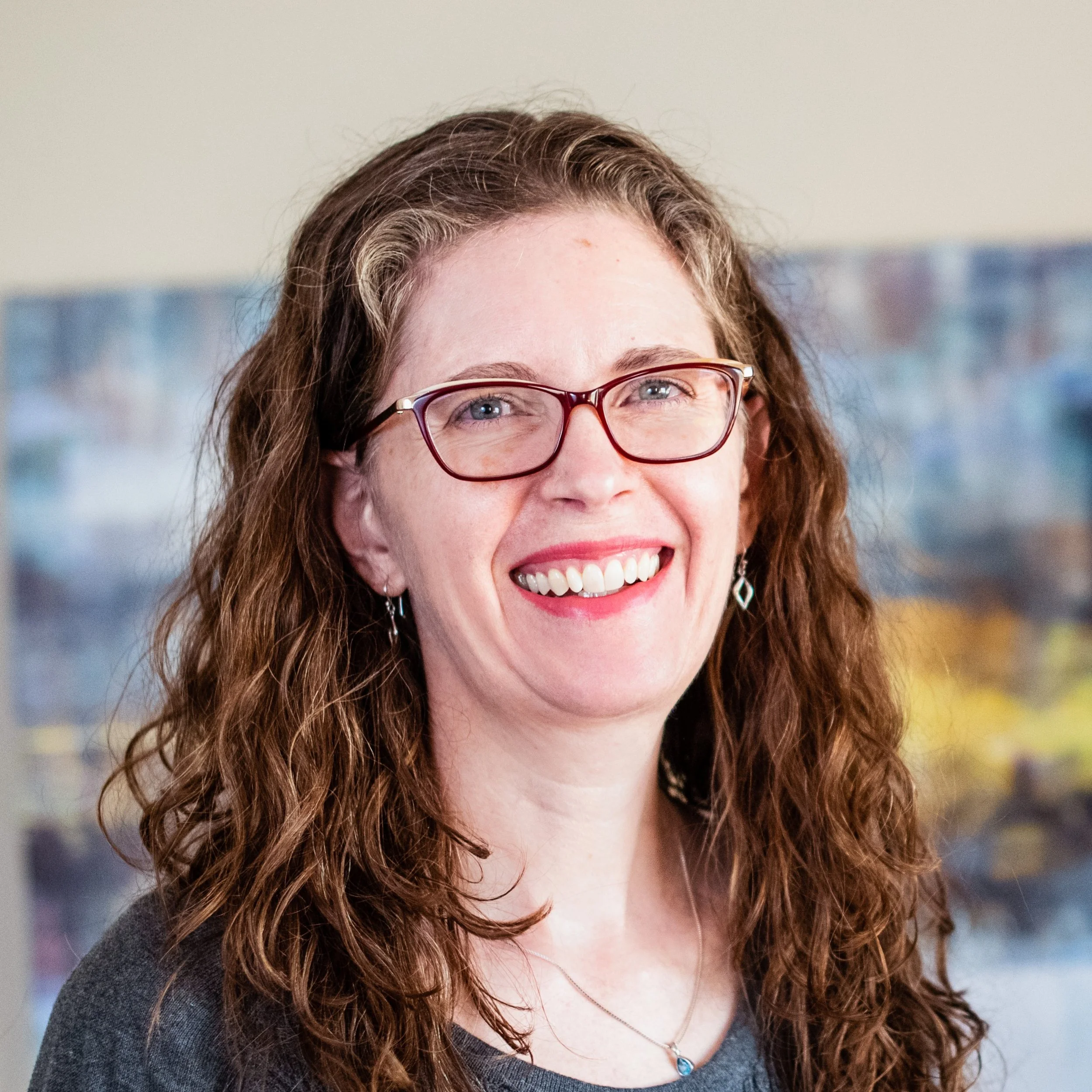 Close-up of a smiling woman with curly brown hair, wearing glasses, a necklace with a blue pendant, and a gray top. Background is blurred.