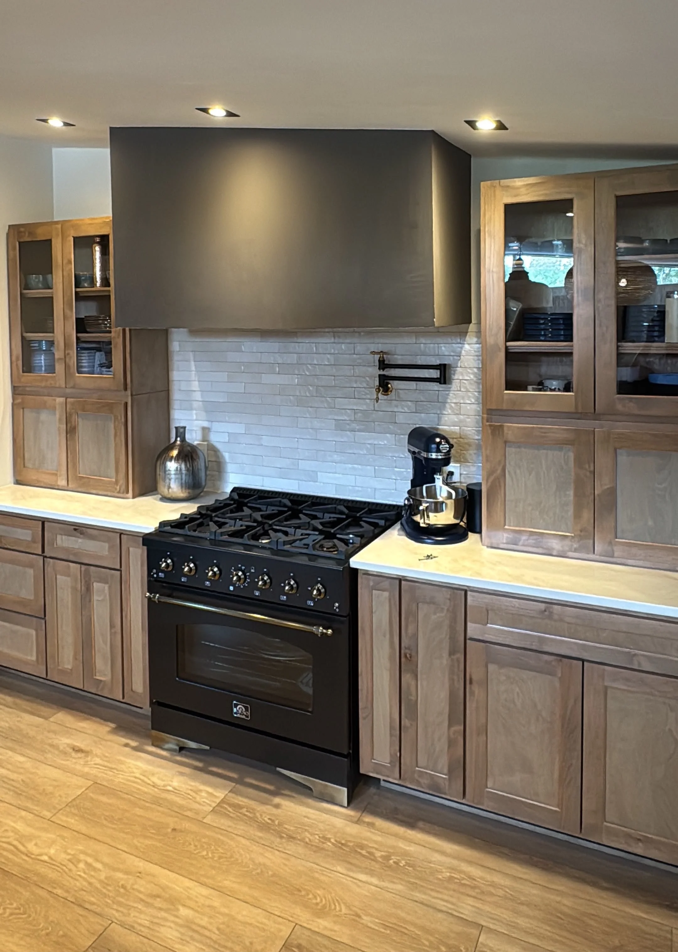 Kitchen with wooden cabinets, a black oven and stove, a white brick backsplash, and a black range hood. There are kitchen appliances, including a silver vase and a stand mixer, on the countertops, and recessed lighting in the ceiling.