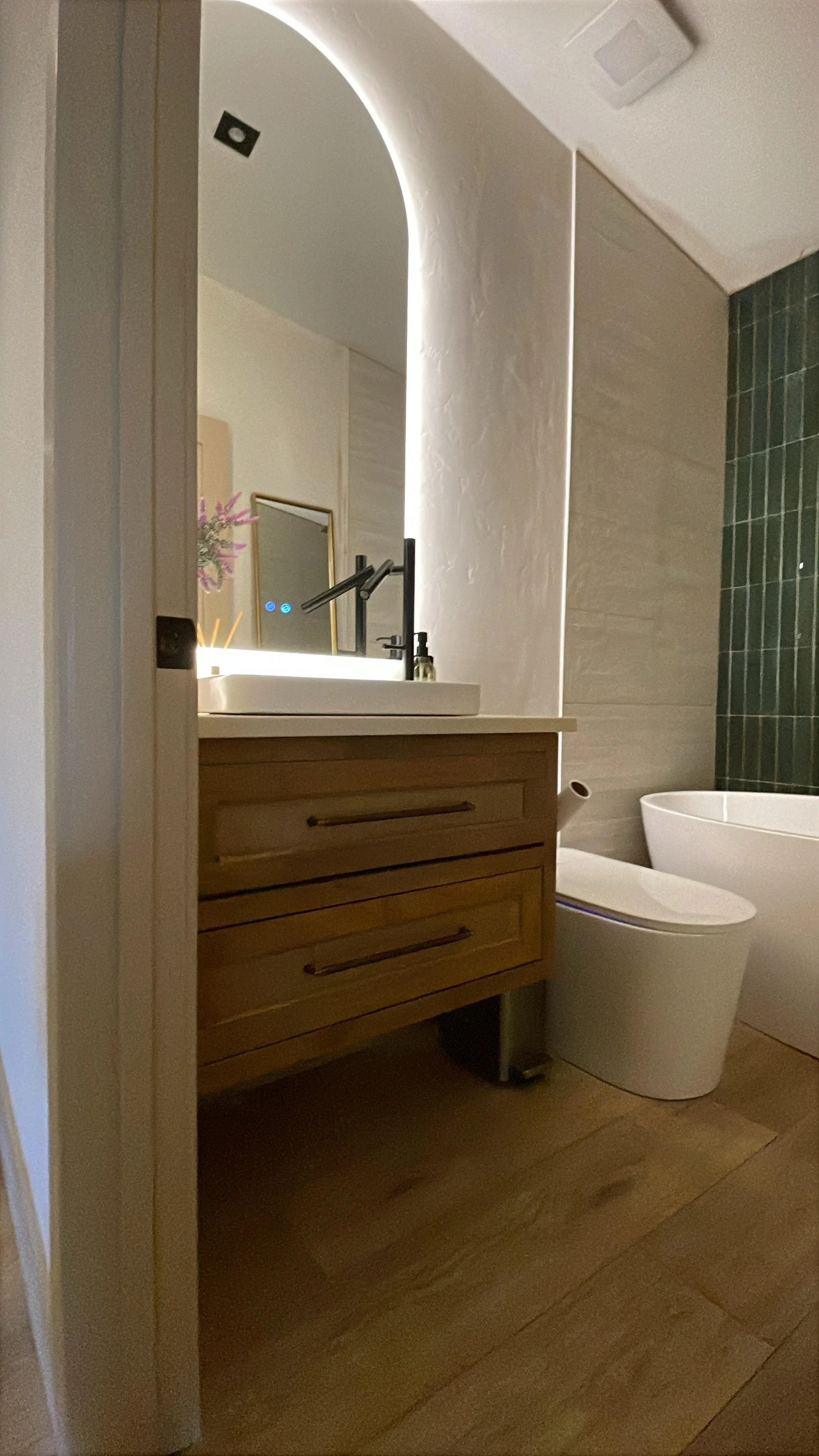 A modern bathroom featuring a wooden vanity with a white countertop and black faucet, a mirror reflecting a purple flower, a white toilet, and a partially visible shower area with dark green tiles.