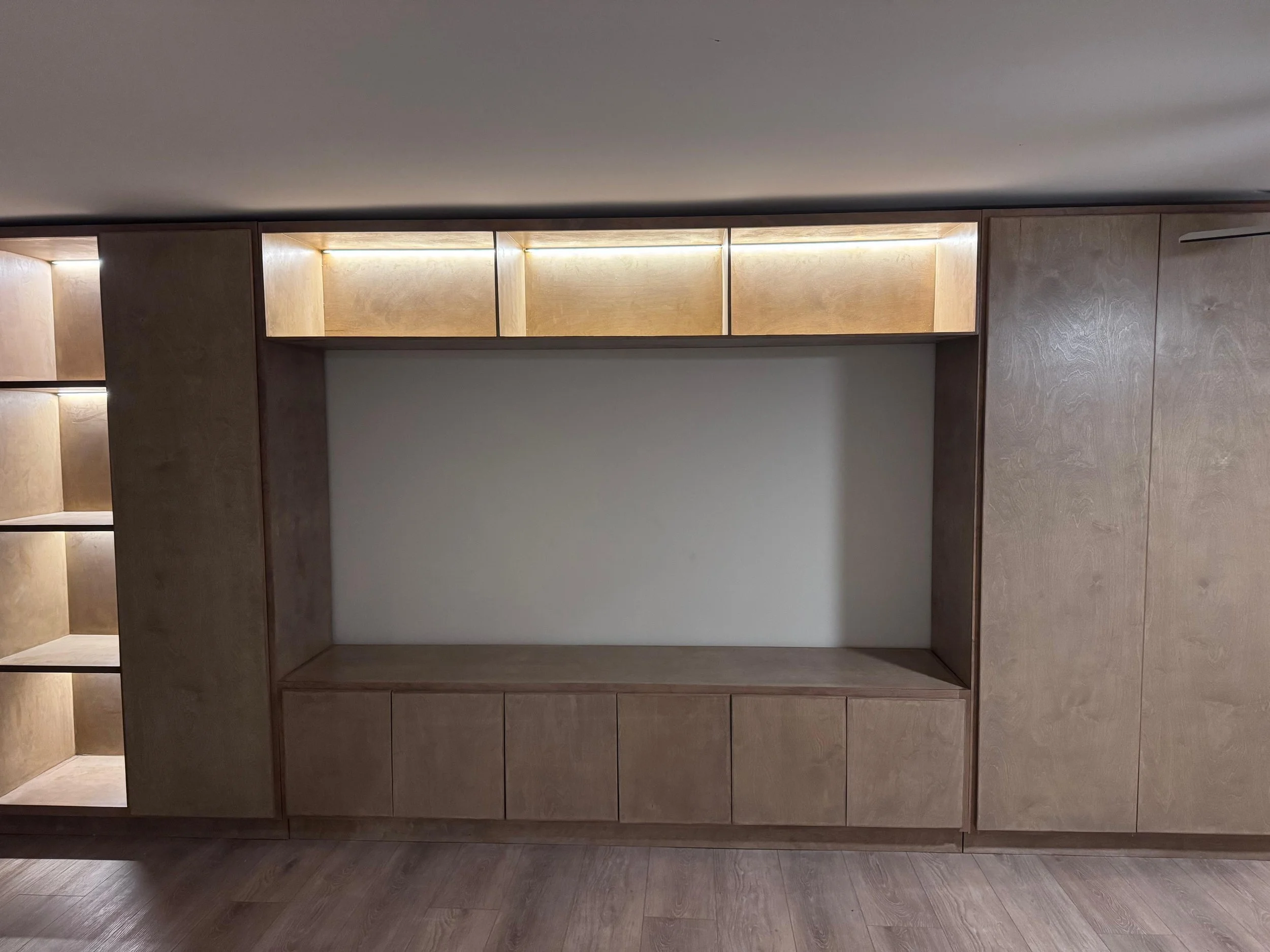 A modern built-in wooden cabinet with open shelves and closed storage compartments, illuminated with warm lighting, set against a wall with wood flooring.