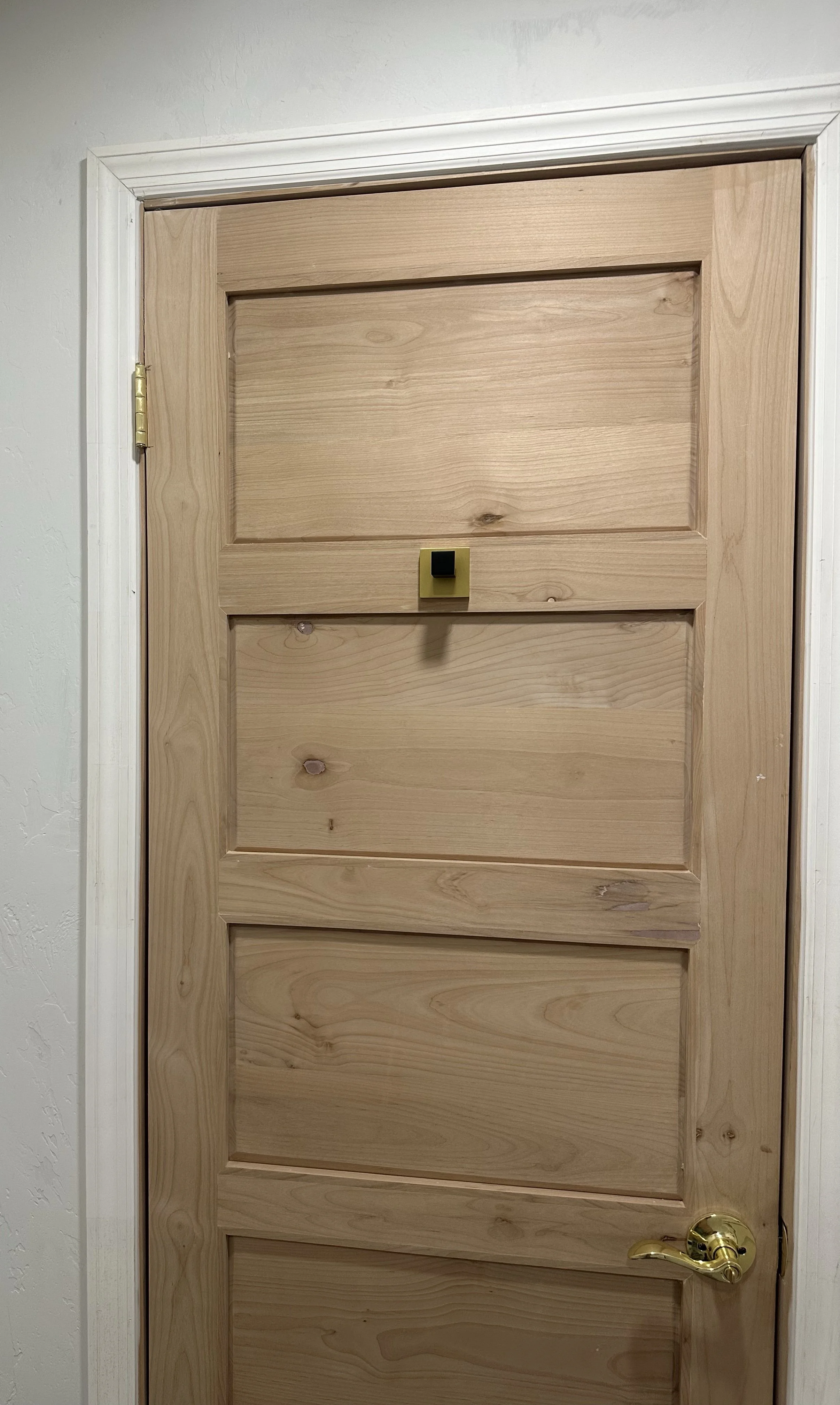 Unfinished wooden door with a small square black latch and a gold doorknob, surrounded by white trim on a white wall.