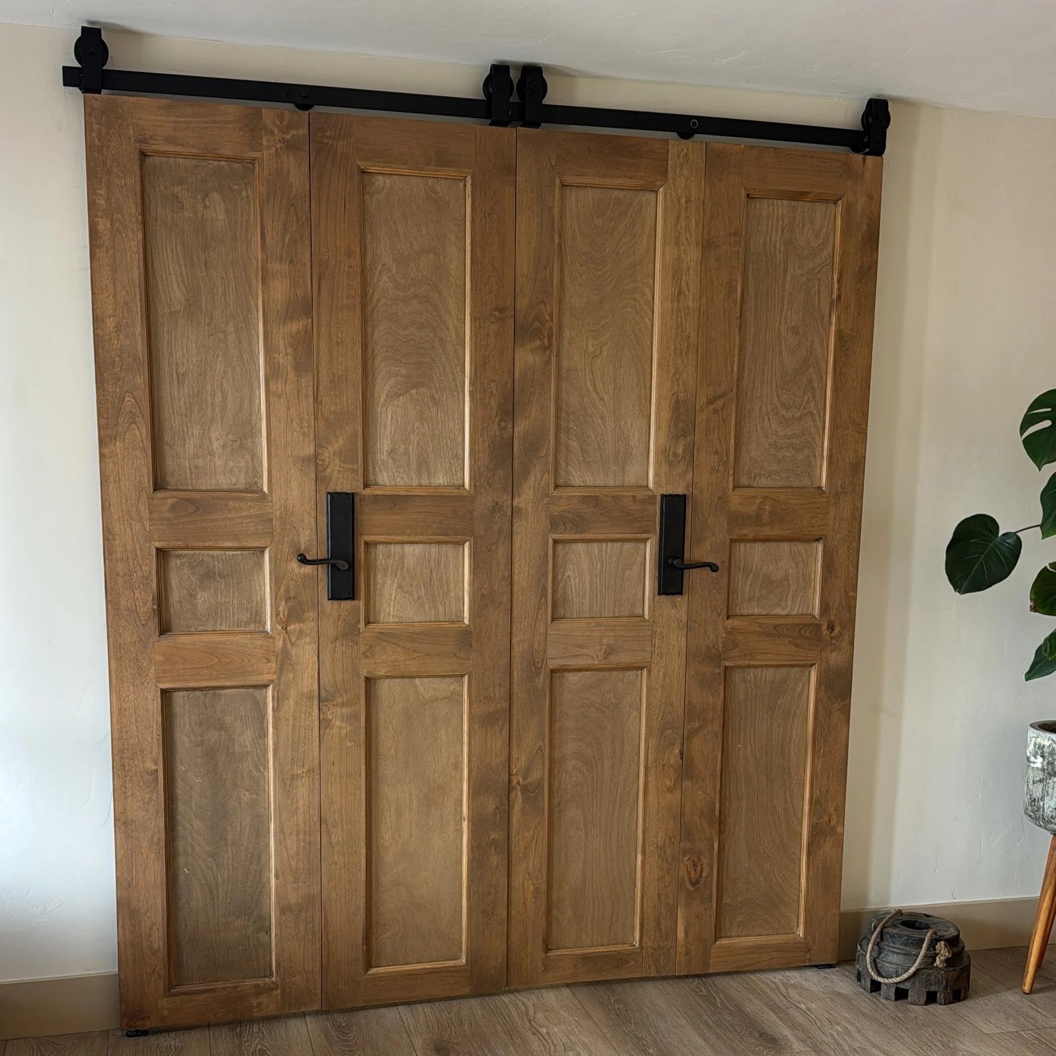 Wooden sliding barn door with black hardware on a white wall, overlooking a hardwood floor and some decorative items like a basket and a plant.