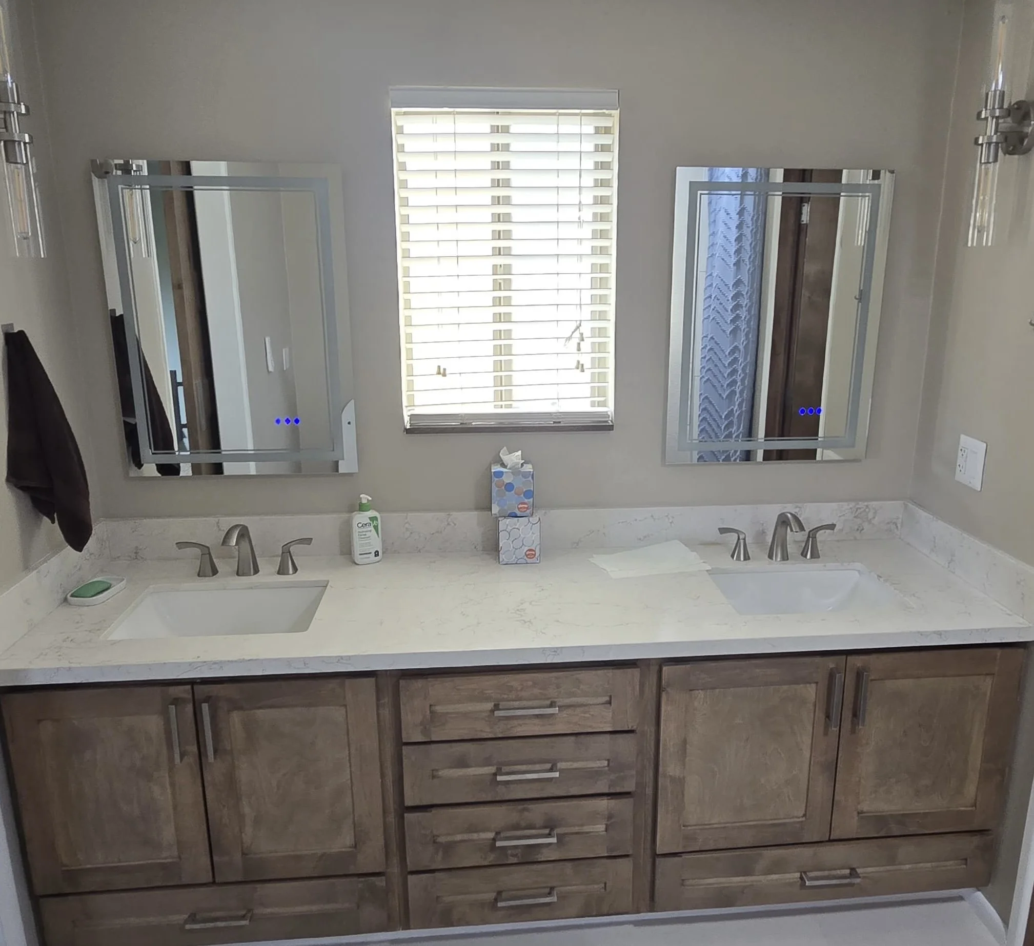 Bathroom vanity with a white countertop, a built-in sink, two faucets, two mirrors, a window with blinds, a tissue box, a soap dispenser, and a bottle of hand sanitizer.