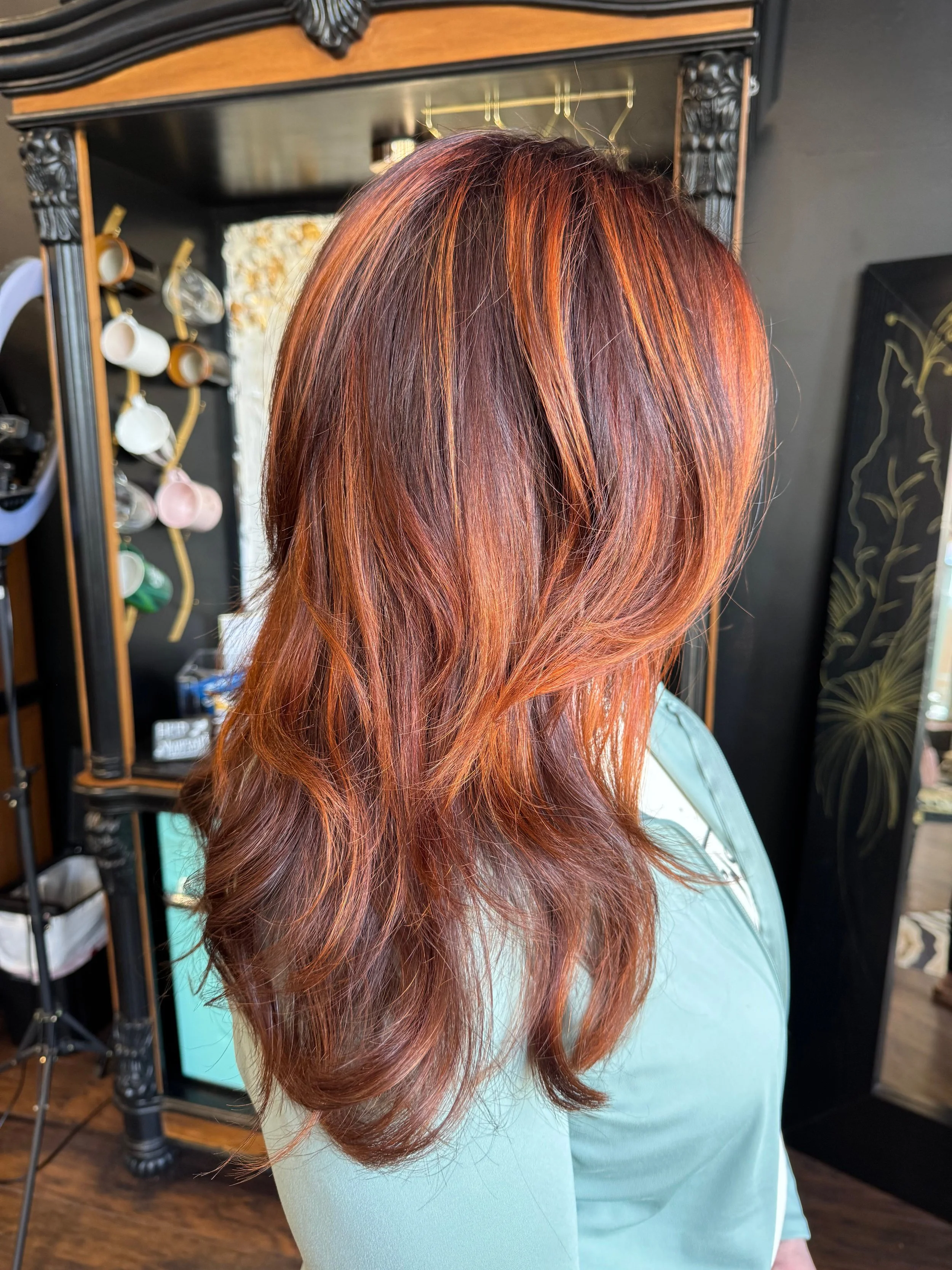 A woman with red auburn hair styled in loose waves, wearing a light blue top, standing in a hair salon.
