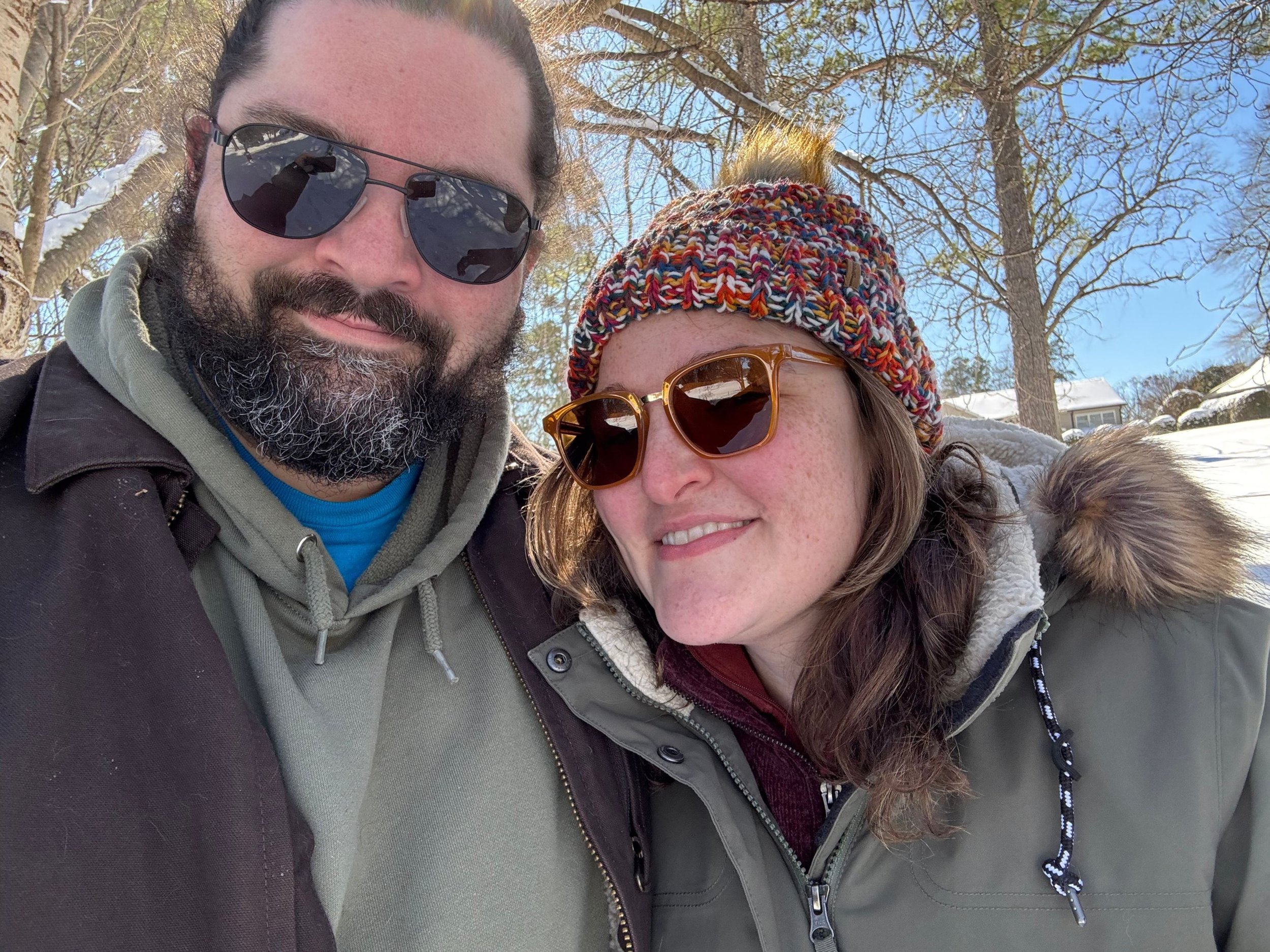 Close-up of two people, a man with dark hair, a beard, and sunglasses, and a woman with long hair, sunglasses, and a colorful knit hat, taking a selfie outdoors on a snowy day with leafless trees and a house in the background.