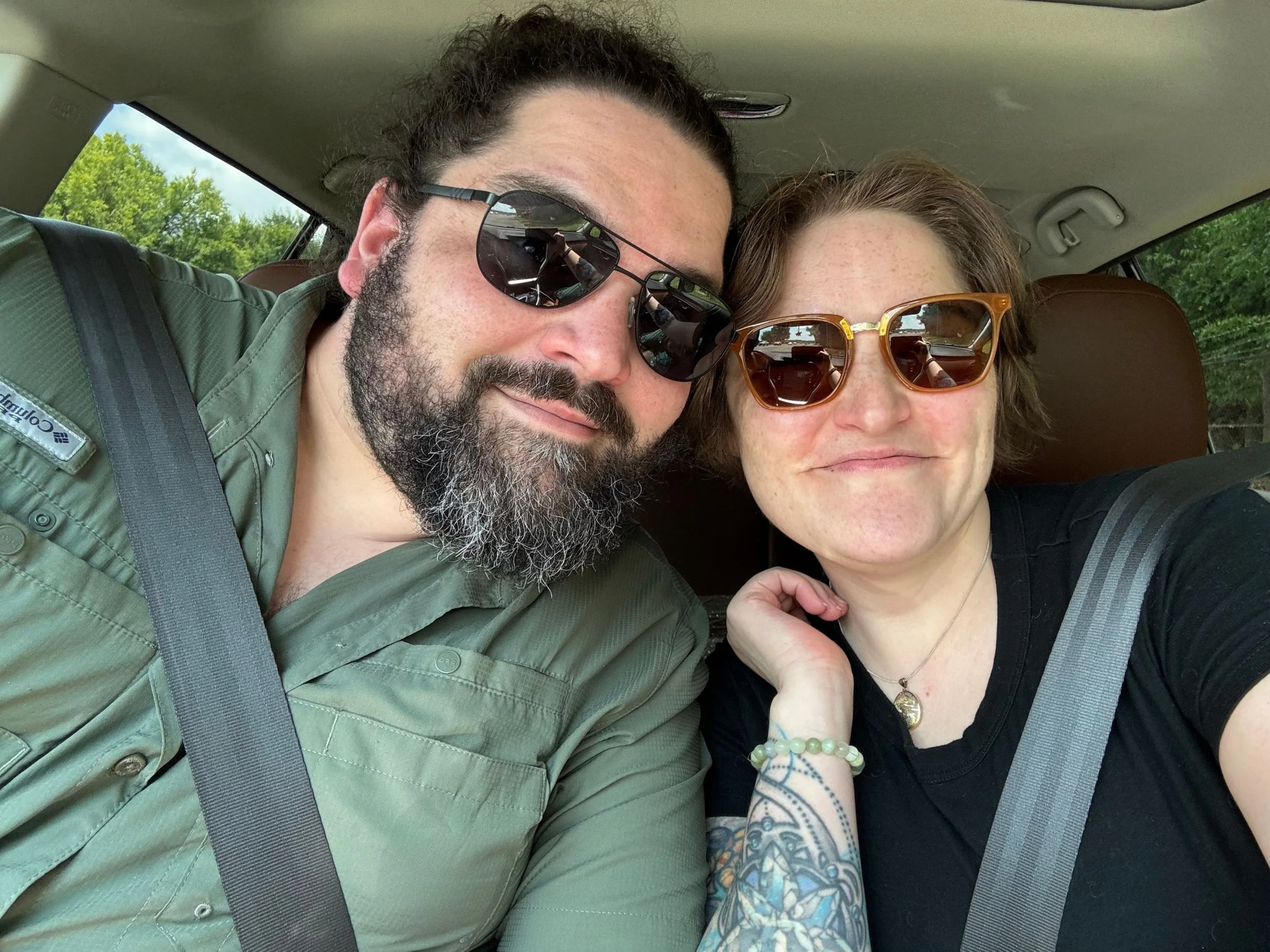 A man and woman sitting in a car, close together, wearing sunglasses, smiling at the camera.