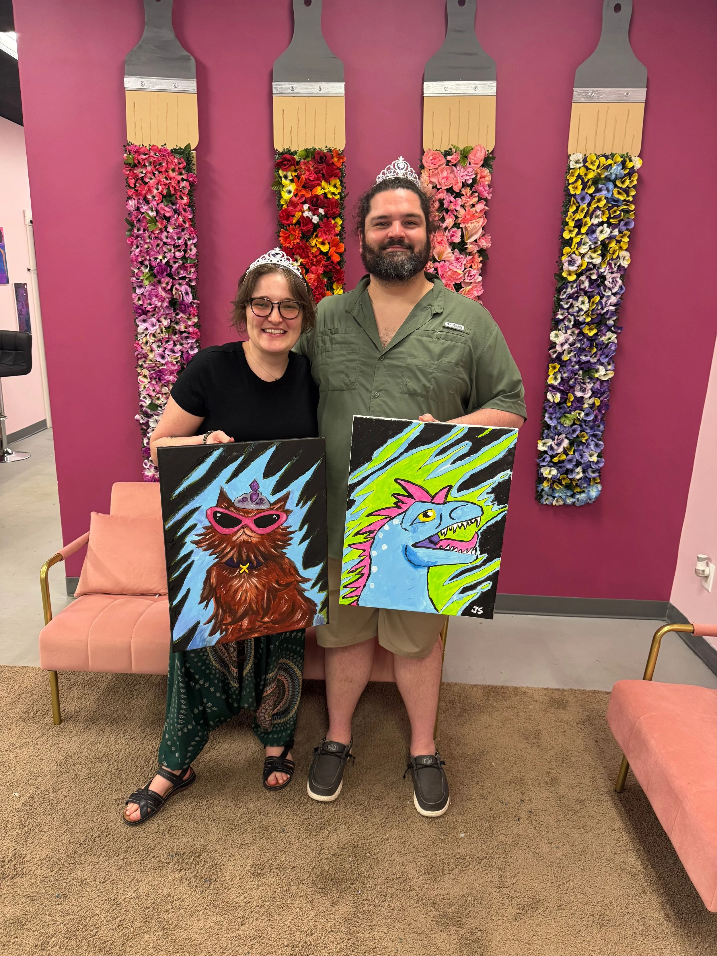 Two people, a woman and a man, standing side by side in front of a pink wall decorated with large paintbrushes and flowers. Both are wearing tiaras and holding colorful paintings of animals. The woman has short brown hair, glasses, and is smiling, wh