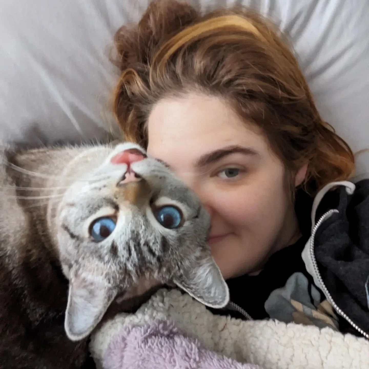 A woman with reddish hair lying close to a kitten with blue eyes, both resting on a soft blanket.