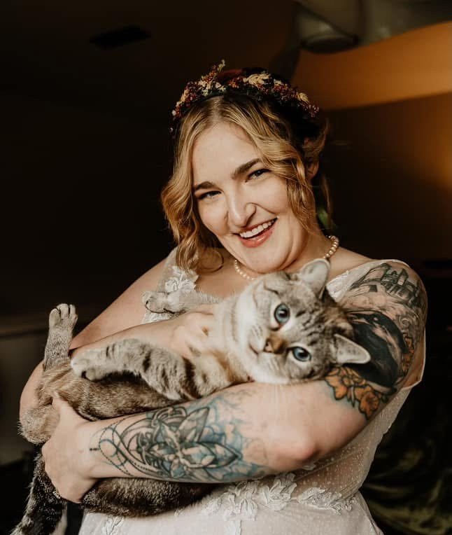 A woman with blonde hair, wearing a floral headband and pearl necklace, holding a gray tabby cat with blue eyes, smiling with a happy expression.