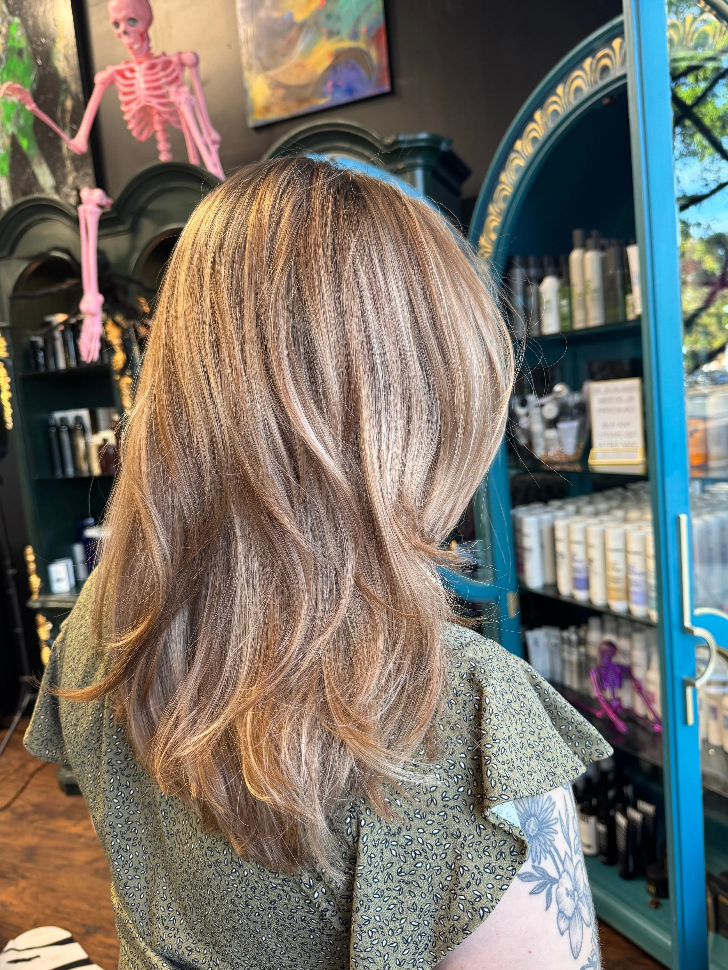 Woman with long, wavy blonde hair seen from behind in a hair salon, holding a patterned purse, with a blue cabinet filled with hair products and a pink skeleton decoration hanging on the wall in the background.