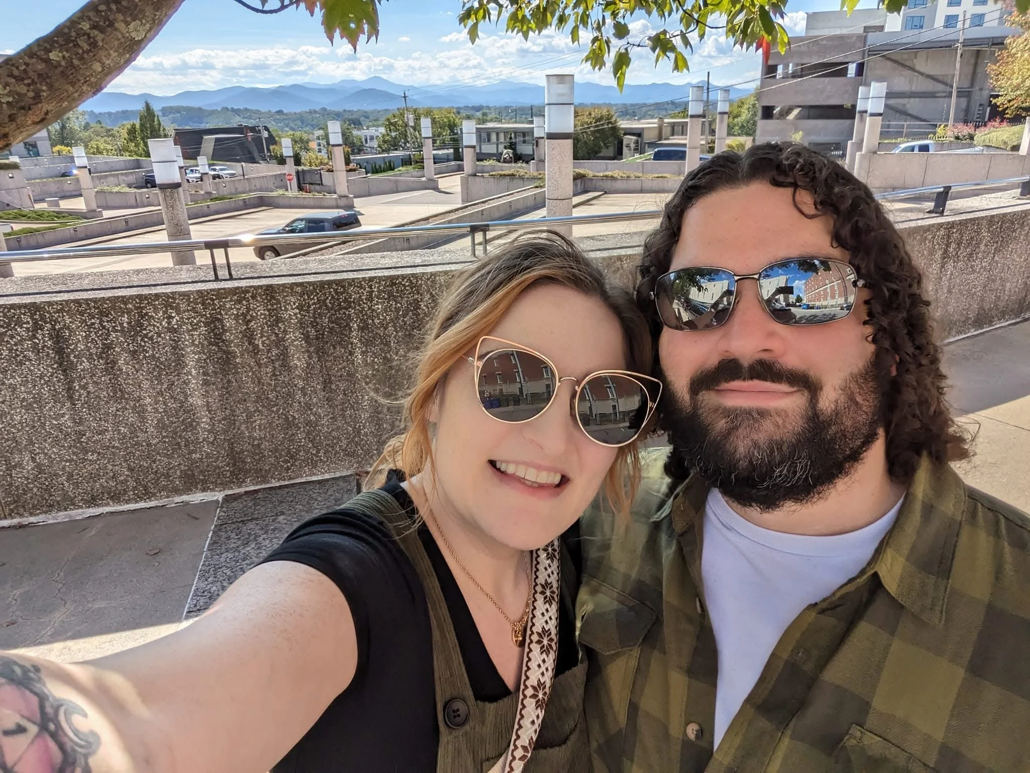Close-up of two people taking a selfie outdoors on a sunny day, with a background featuring a parking lot, trees, buildings, mountains, and a partly cloudy sky.