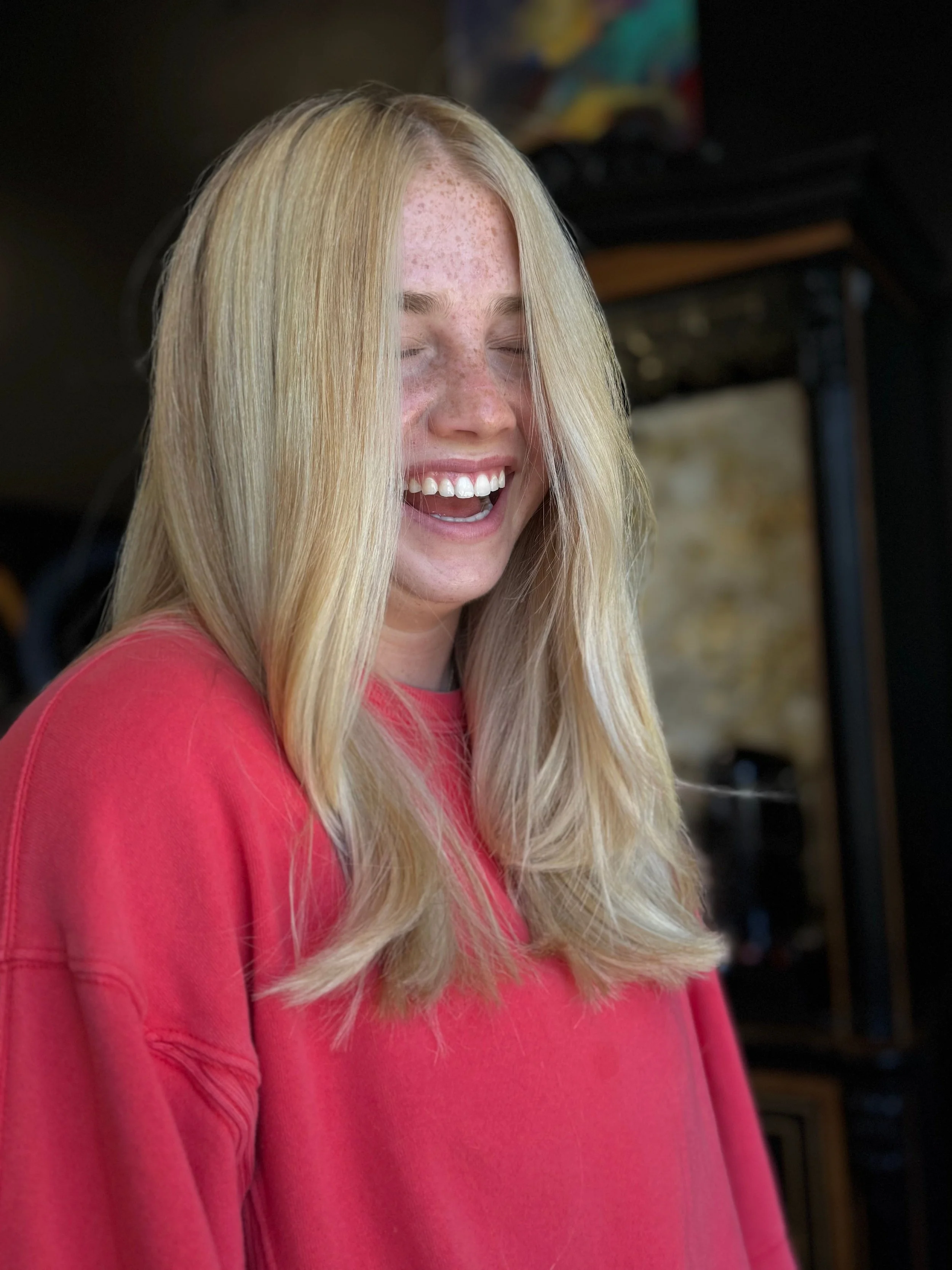A young woman with long, blonde hair, freckles, and closed eyes, wearing a pink top, appears to be happy or enjoying a moment, with a blurred background.