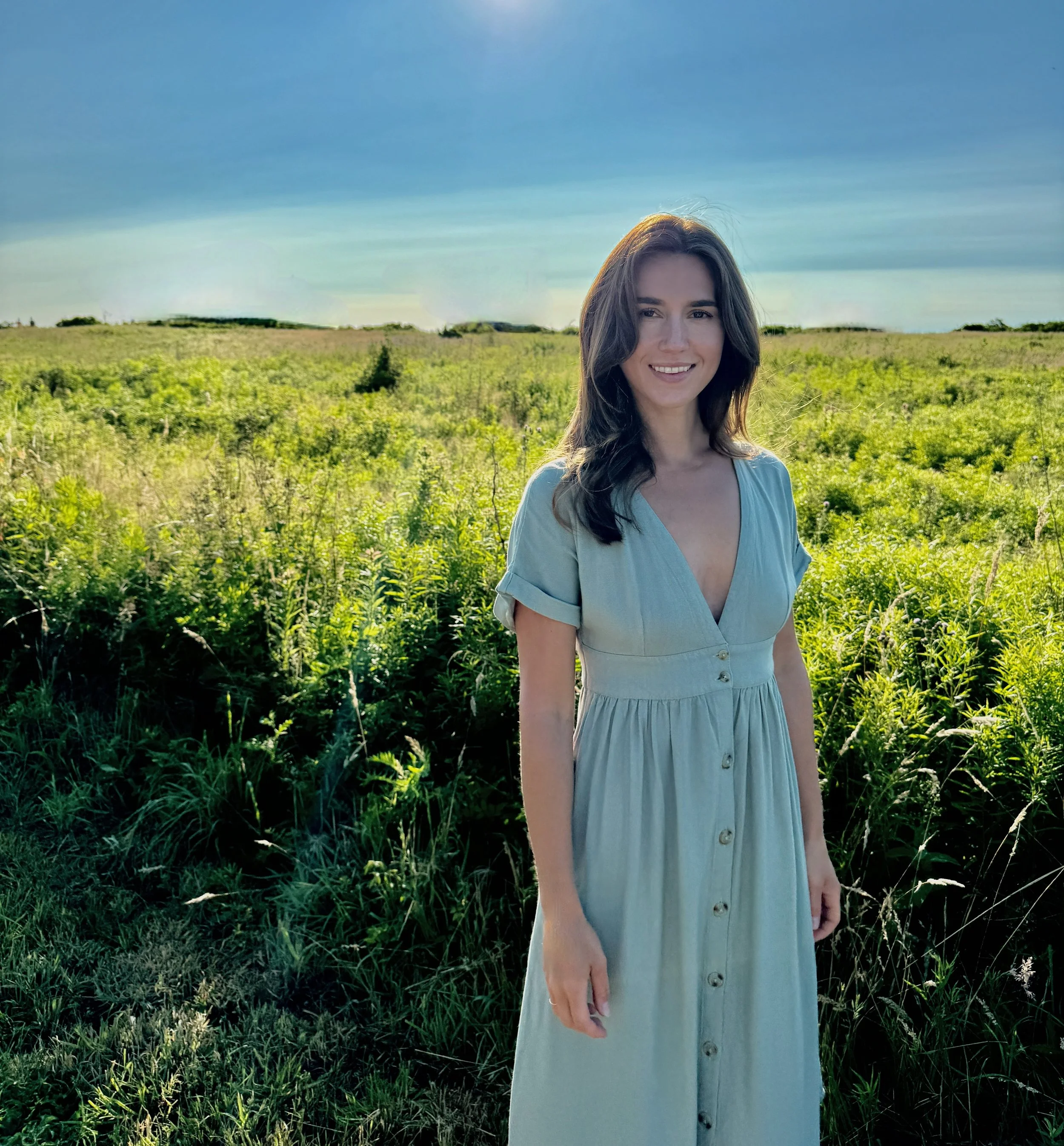 A young woman with long dark hair smiling outdoors in a green field under a blue sky with a few clouds.
