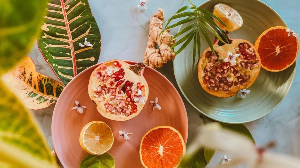 Two colorful bowls contain halved oranges with pomegranate seeds and small white flowers, surrounded by tropical leaves, ginger root, lemon slices, and a small insect on a light blue surface.