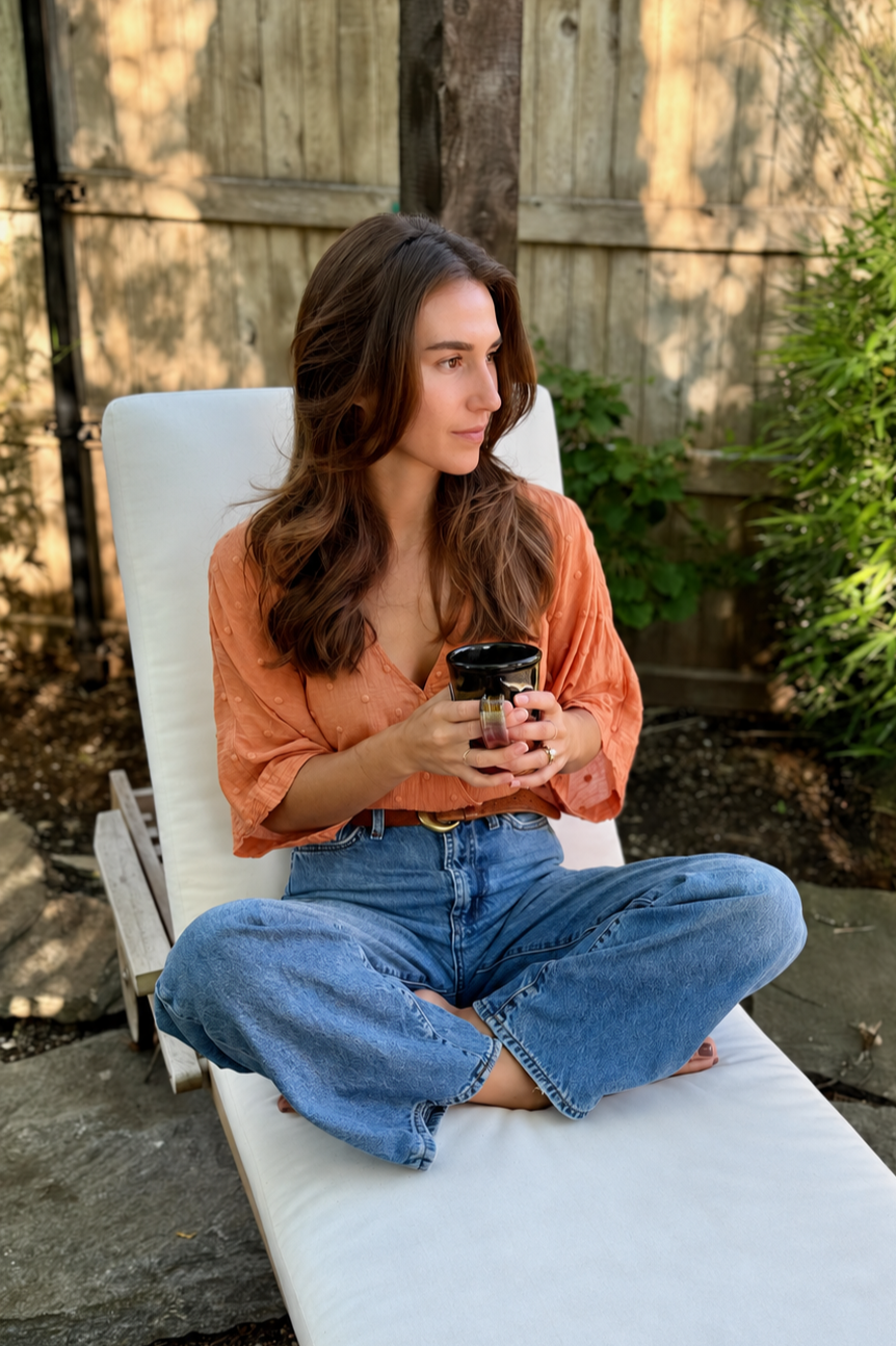 A young woman sitting cross-legged on a lounge chair outdoors, holding a black mug, wearing an orange blouse and blue jeans, with a wooden fence and greenery in the background.