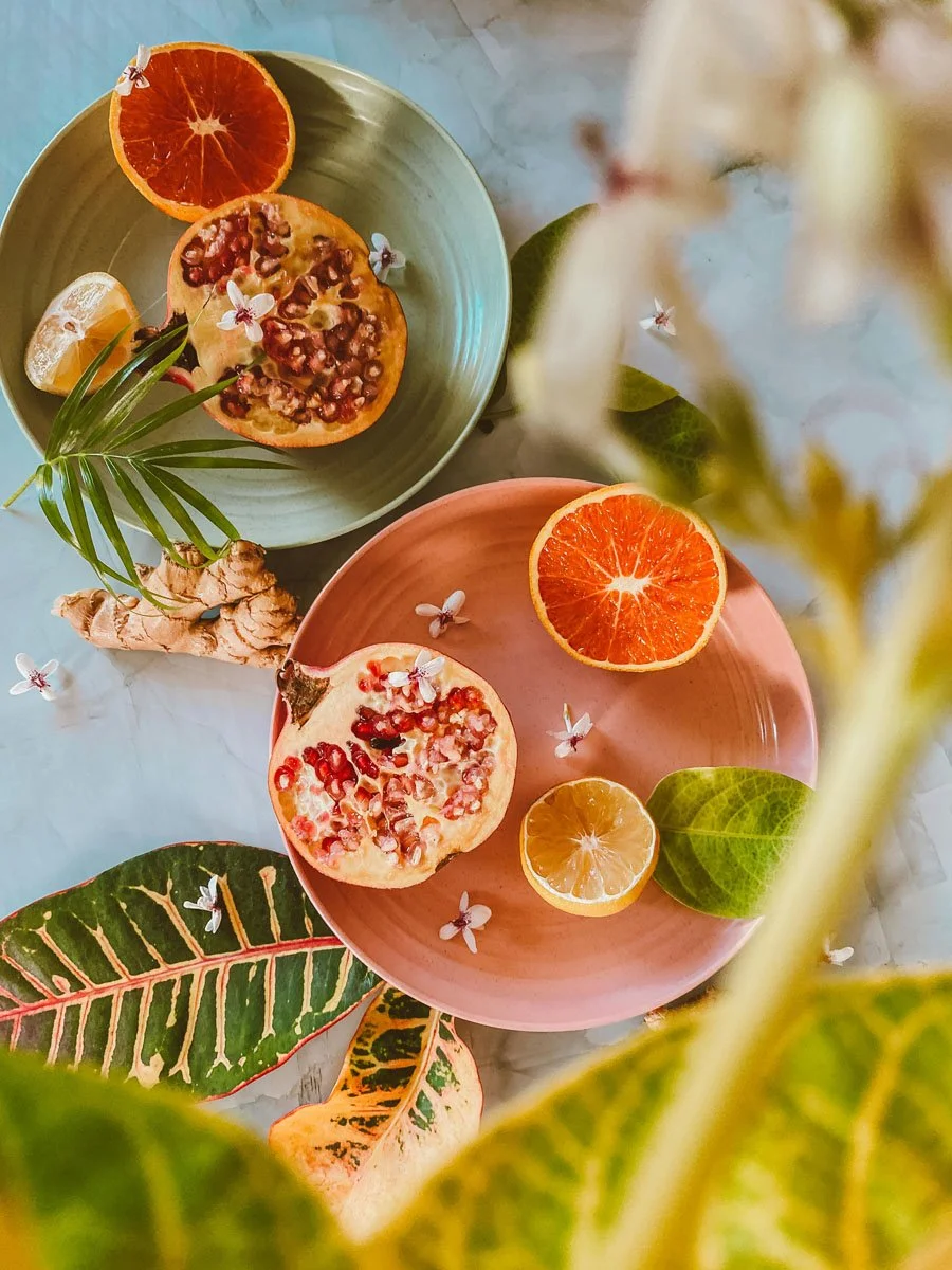 Top-down view of two plates with pomegranate halves and citrus slices, surrounded by green and patterned leaves, with small white flowers scattered around.