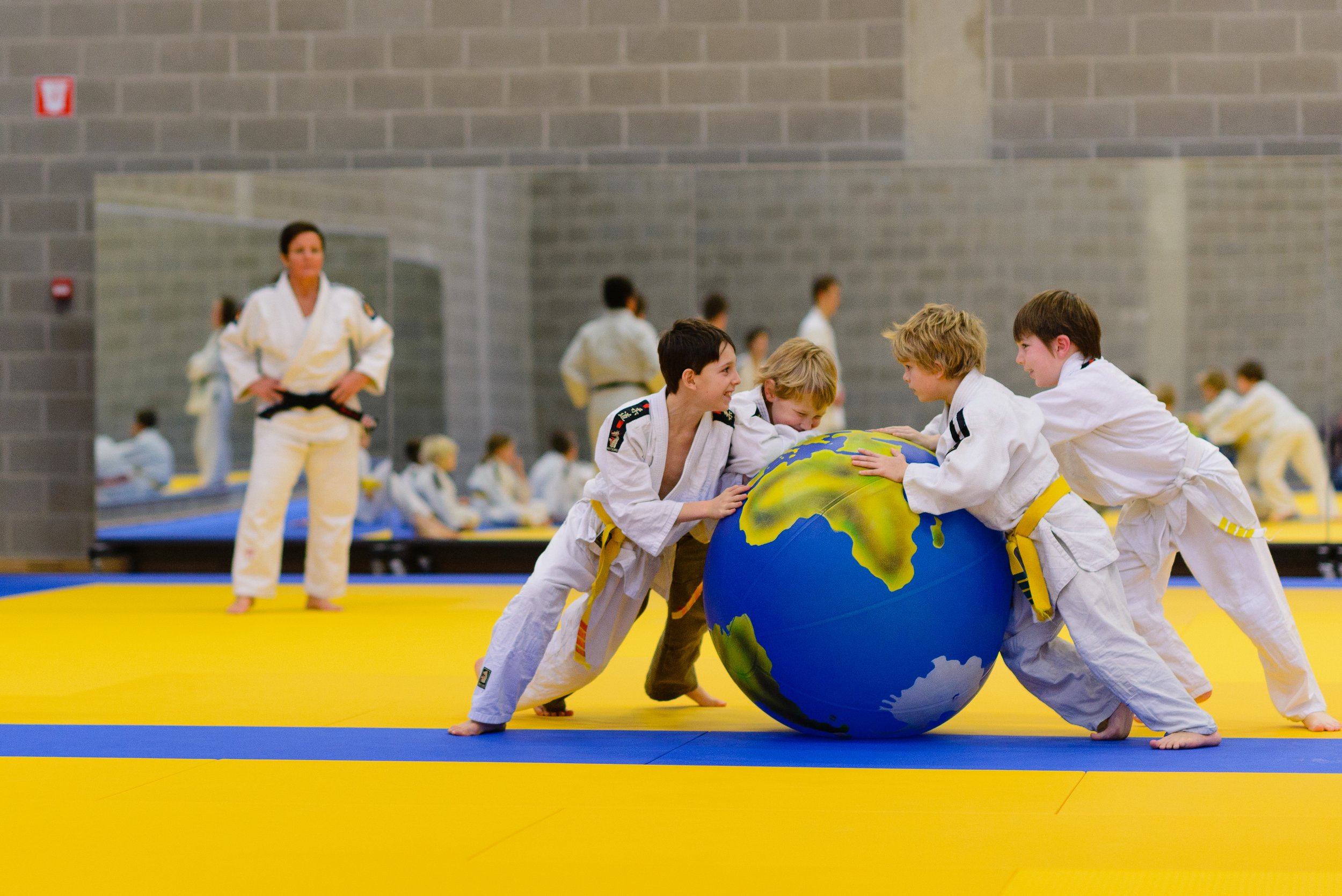 Young martial arts students in gi uniforms participate in a group activity with a large globe, on a yellow and blue mat, in a martial arts training hall. An instructor observes in the background.