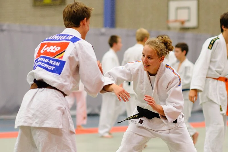 Two judo practitioners, a young man and a young woman, engaging in a sparring match on a dojo mat, with others practicing in the background.
