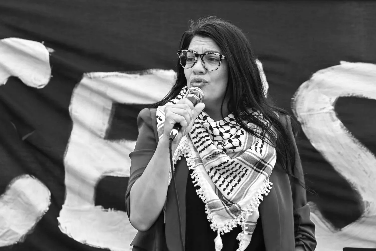 A woman with dark hair, wearing glasses, a patterned bandana, and a blazer, speaking into a microphone at an outdoor event.