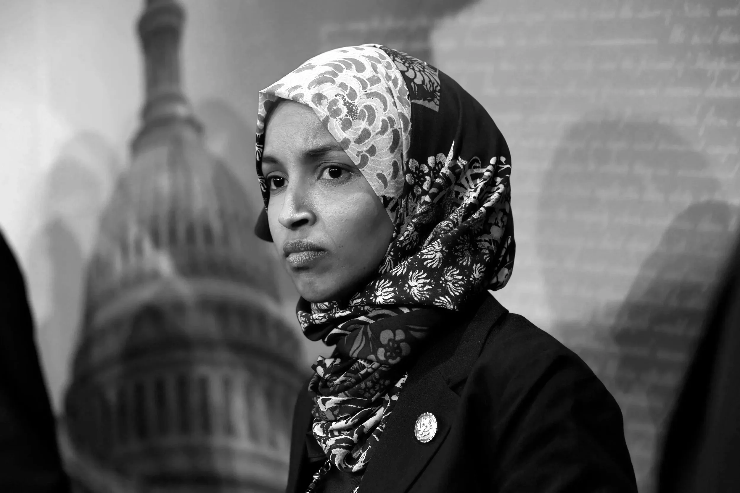 A woman wearing a patterned headscarf and a dark jacket with a badge, looking to her right. The photo is black and white and taken indoors.