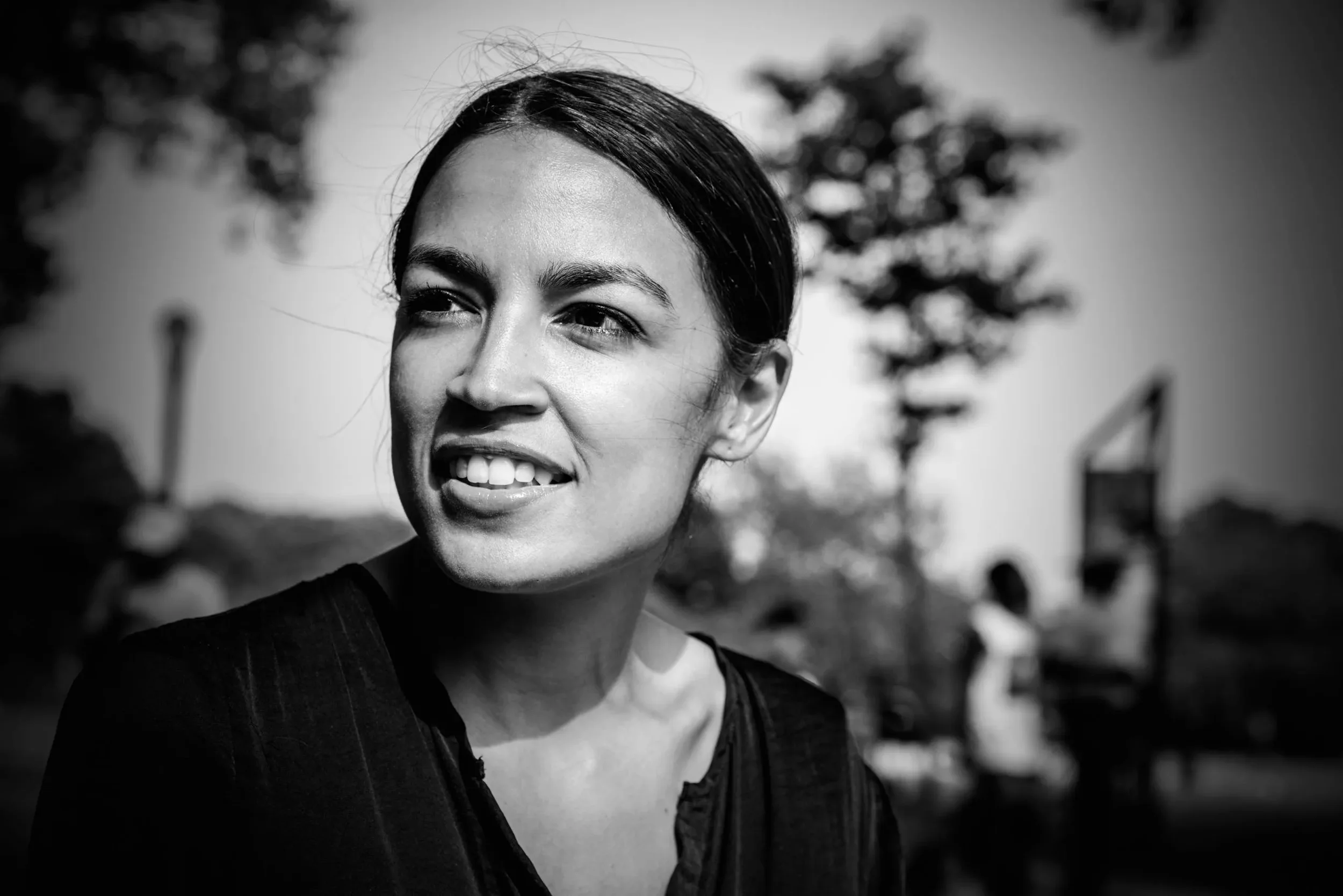 A black and white close-up portrait of a smiling woman with dark hair, outdoors with blurred trees and buildings in the background.