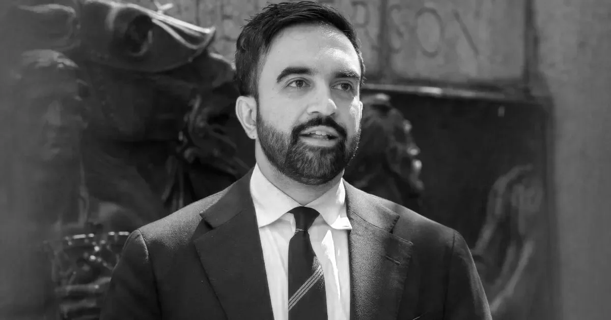 A man with dark hair and beard wearing a suit and tie, standing indoors with graffiti and backpacks in the background.