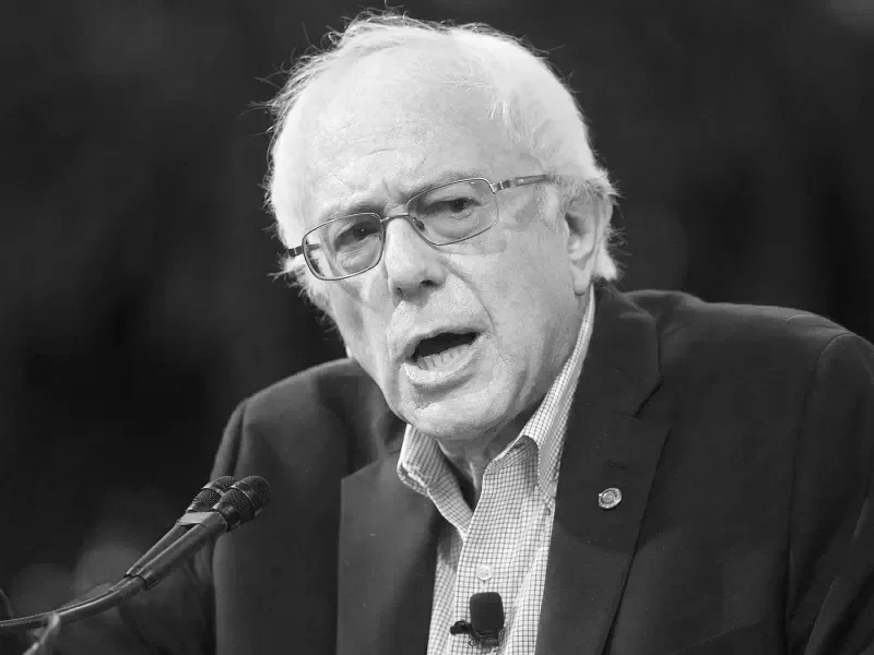 A black-and-white photo of Bernie Sanders speaking into a microphone at a public event or hearing, with a serious expression on his face.