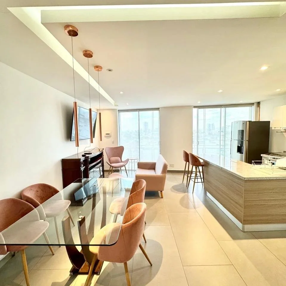 Sala de estar moderna con sillones rosas y beige, mesa de comedor de vidrio, paredes blancas, ventanas grandes con vista de la ciudad, cocina con barra de desayuno y refrigerador negro.