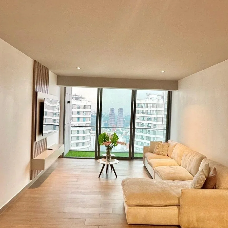 Sala de estar moderna con sillón beige, mesa con flores y vista a edificios altos desde una ventana grande.