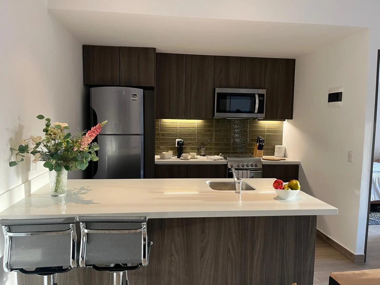 Cocina moderna con isla de encimera blanca, flores en jarra, fruta en tazón, electrodomésticos de acero inoxidable y gabinetes de madera oscuro.