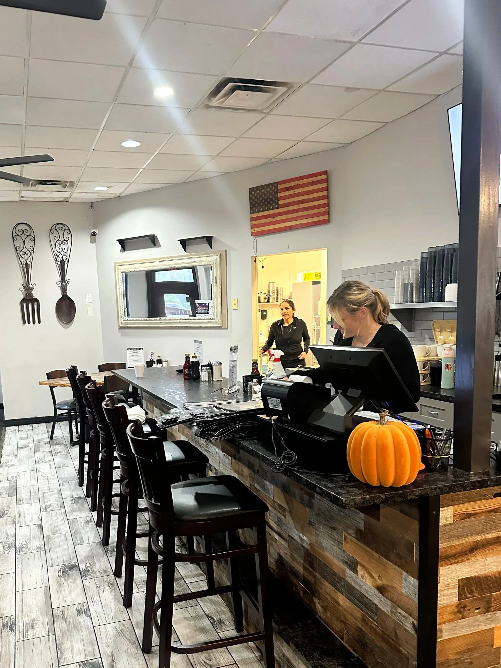Interior of a restaurant or cafe with a long counter, black chairs, a pumpkin decoration, and employees preparing food. There is a wall decor with large forks and spoons, an American flag art piece, and a window.