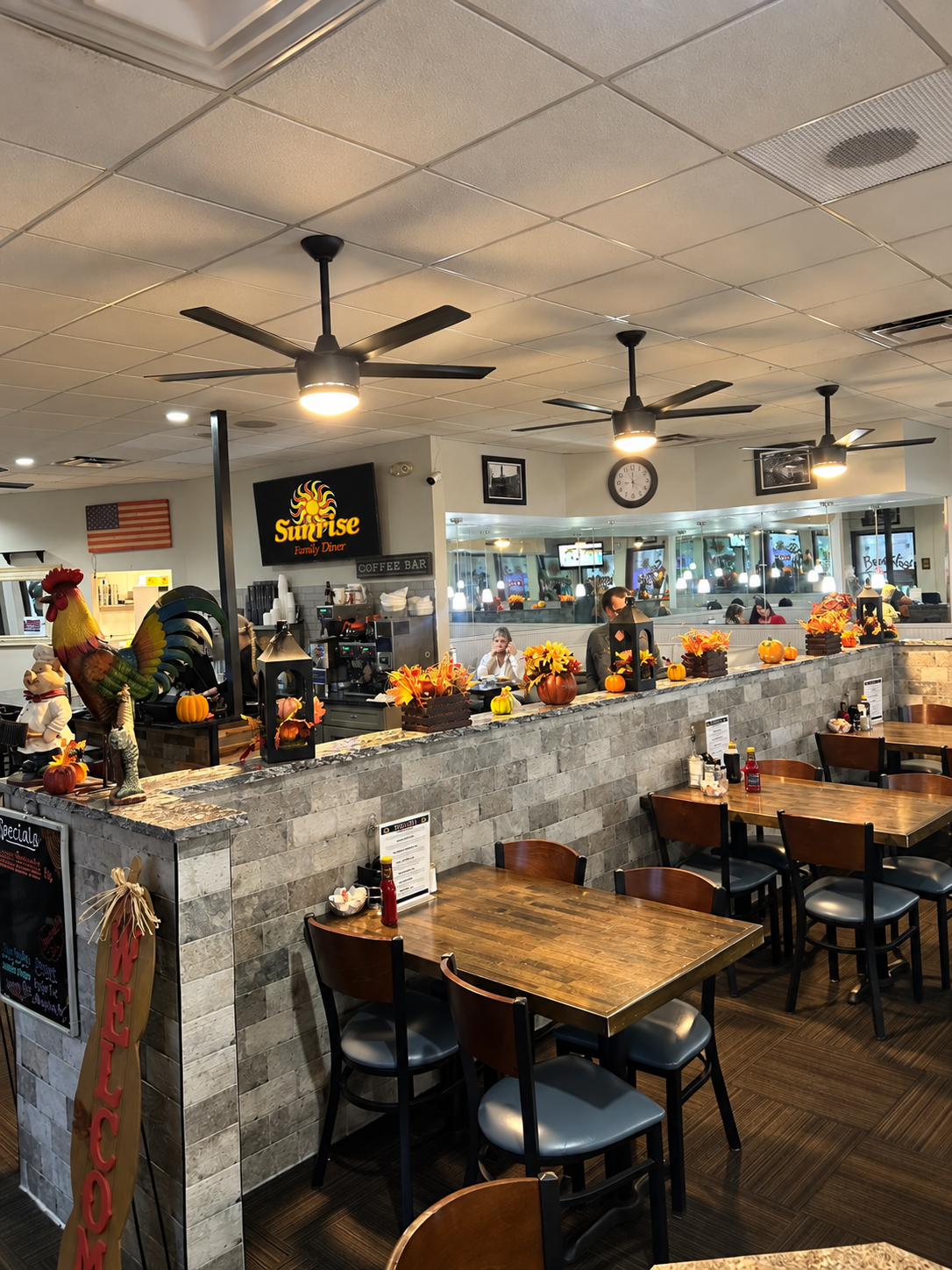 Interior of a family restaurant decorated for autumn with pumpkins and fall flowers, featuring a counter with some staff working behind it, and empty tables with chairs in the dining area.