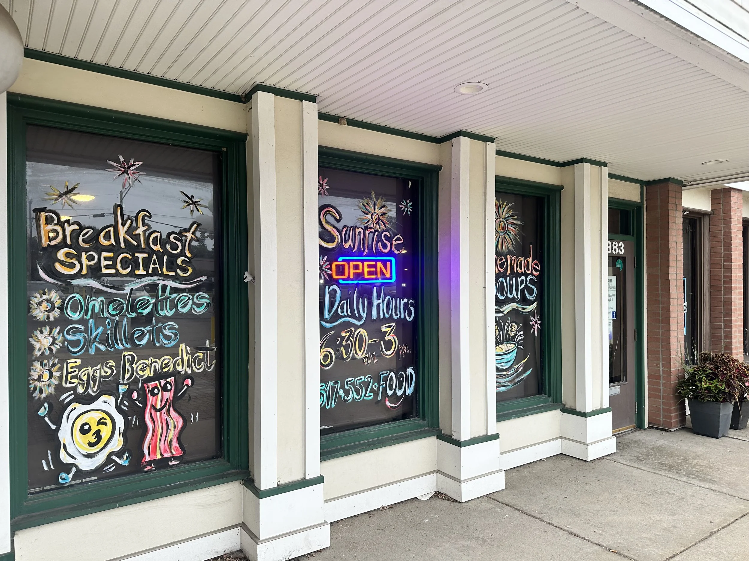 Storefront window with colorful chalk art advertising breakfast specials, omelettes, shillelaghs, and eggs Benedict, along with the store's daily hours and contact information. Neon sign reads 'OPEN' with other colorful signage for sunrise and sunset specials.