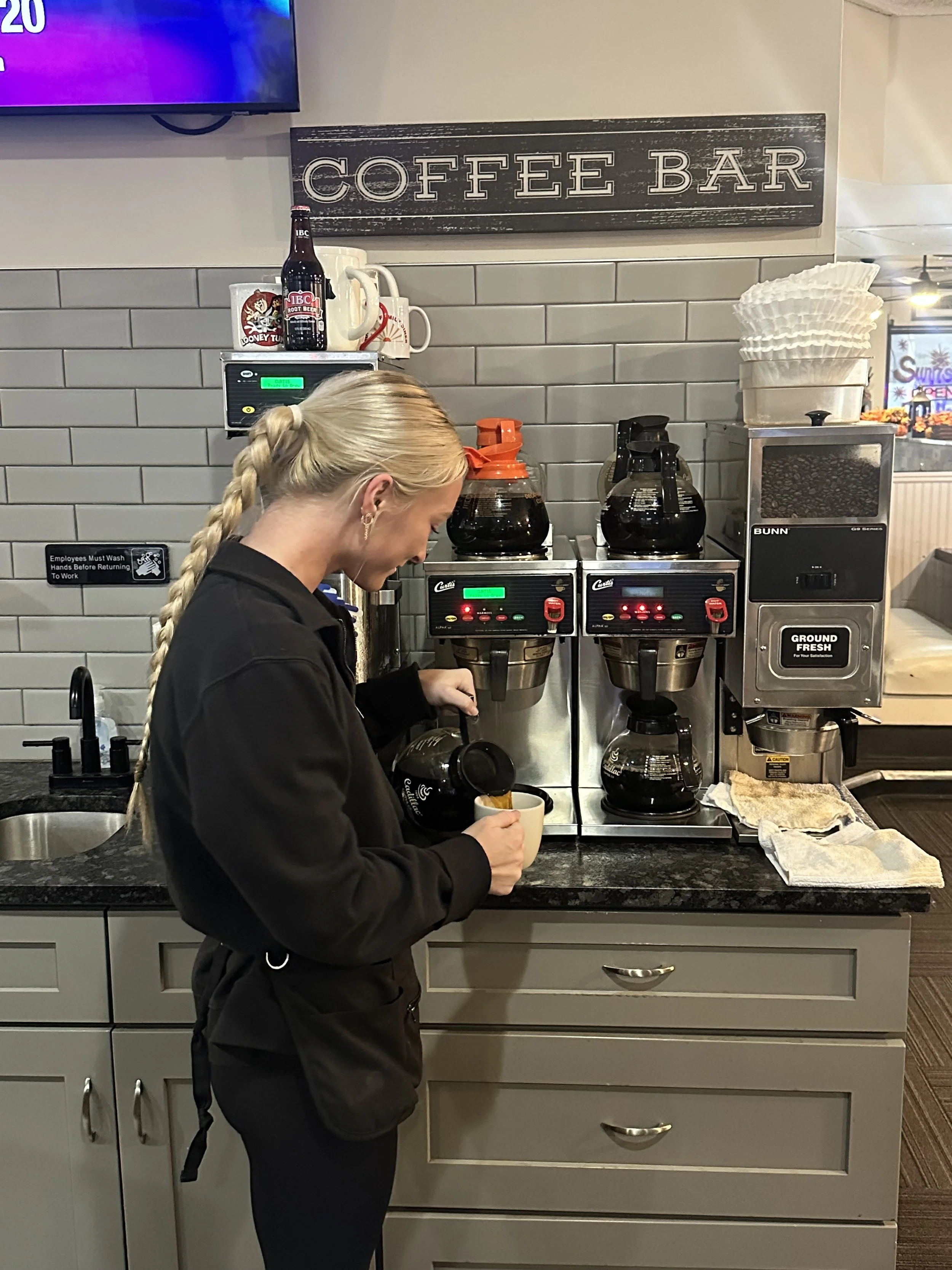 A woman with blonde braided hair, wearing a black jacket, is pouring coffee from a coffee machine into a mug in a coffee shop. There is a sign that says 'Coffee Bar' on the wall behind her, along with coffee brewing equipment and supplies.