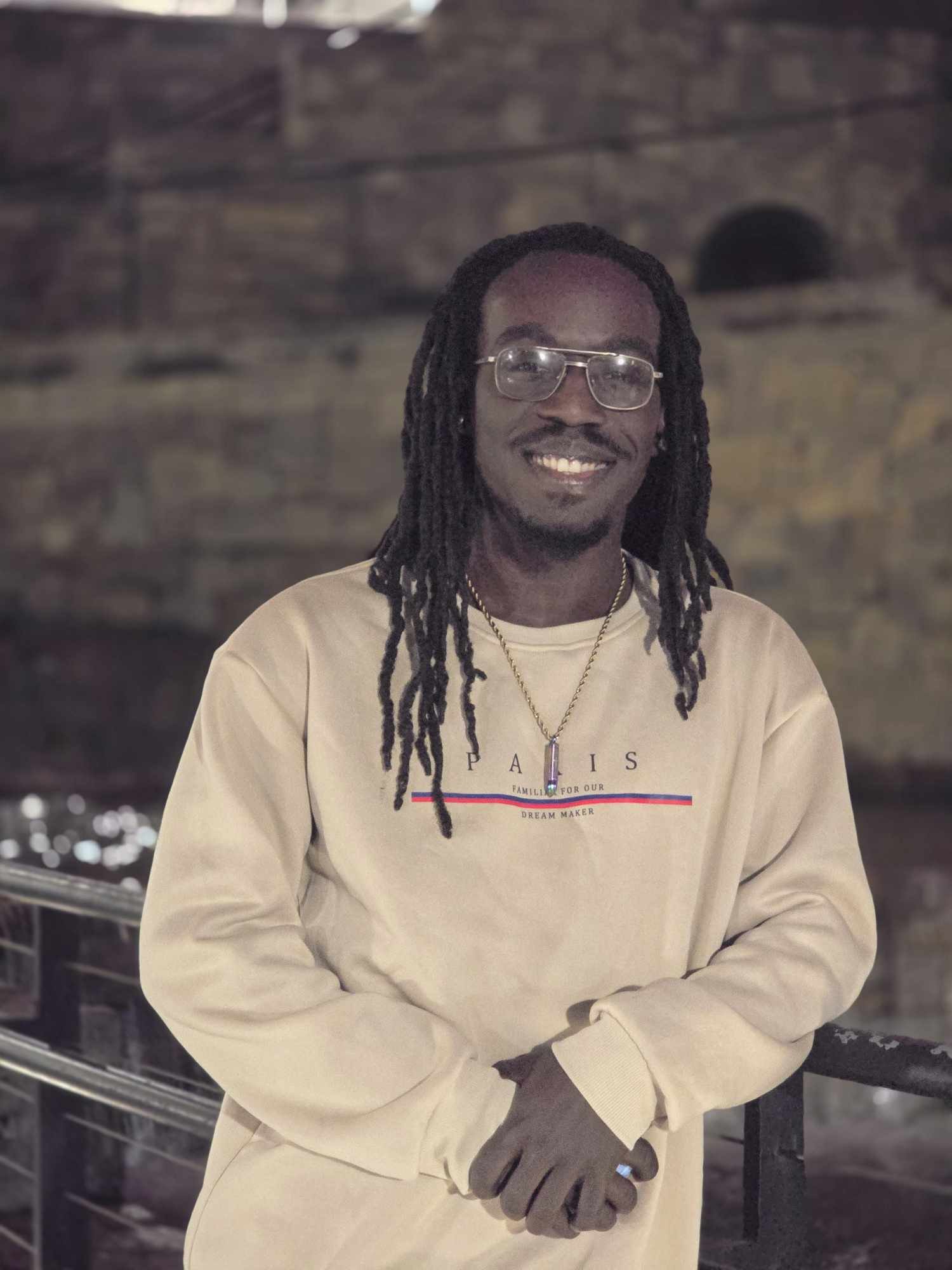 A smiling man with long dreadlocks, wearing glasses and a beige sweatshirt, clasping his hands together in front of him, standing in an indoor setting with a brick wall background.