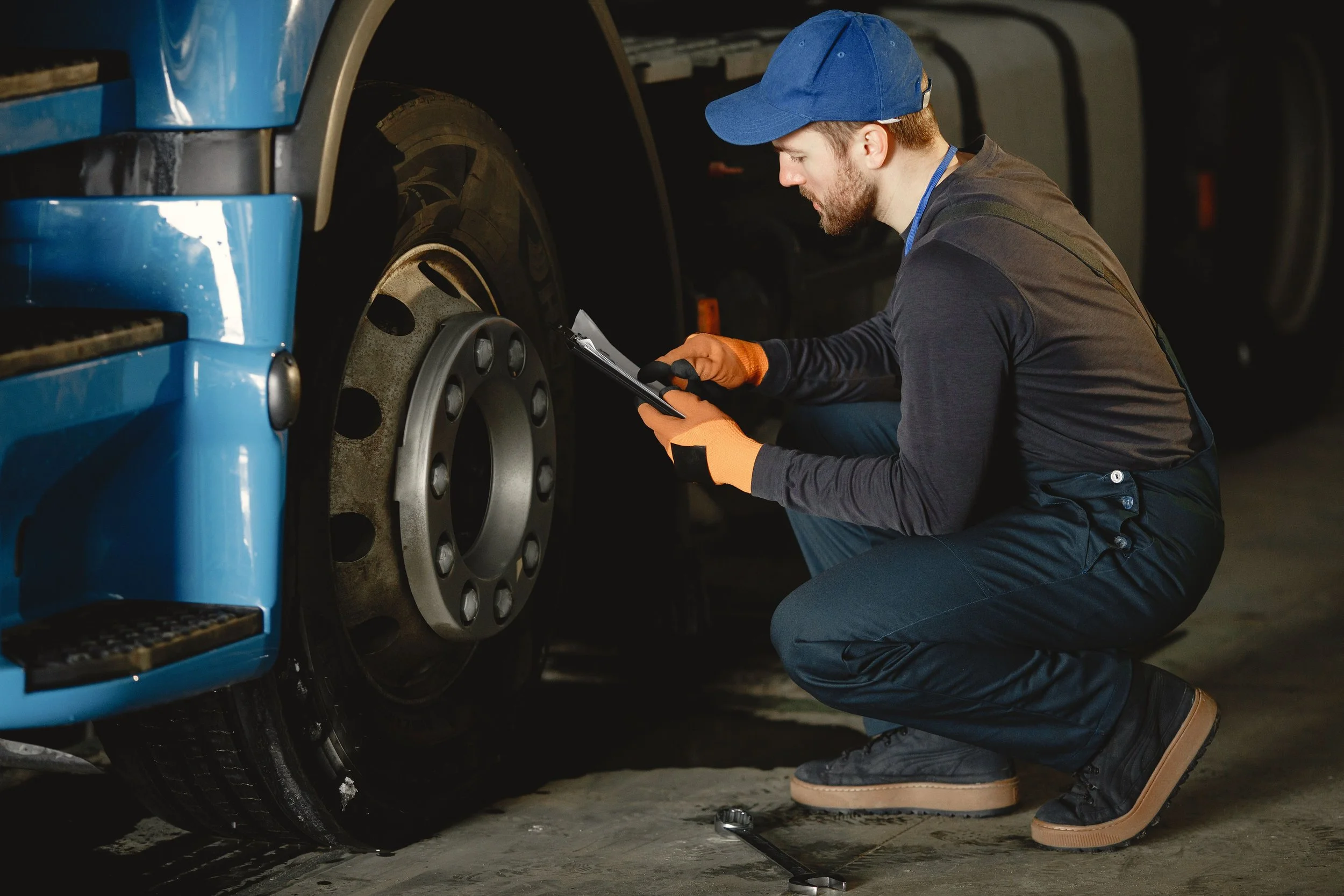 Man kneeling beside a blue truck, inspecting or working on the front tire with a clipboard in hand.