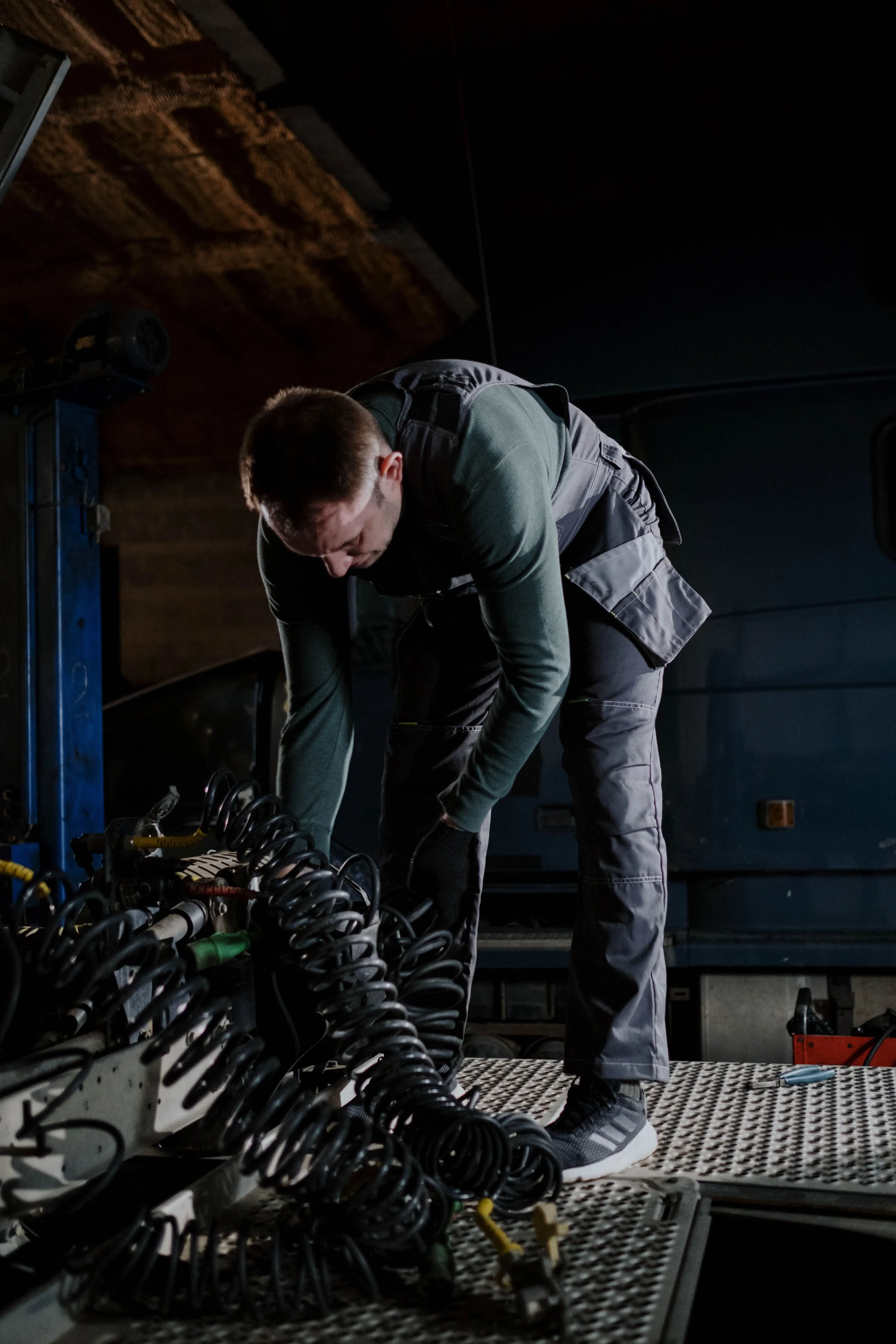 A technician working on electrical equipment in a dimly lit industrial setting, bending over with organized cables and tools around him.