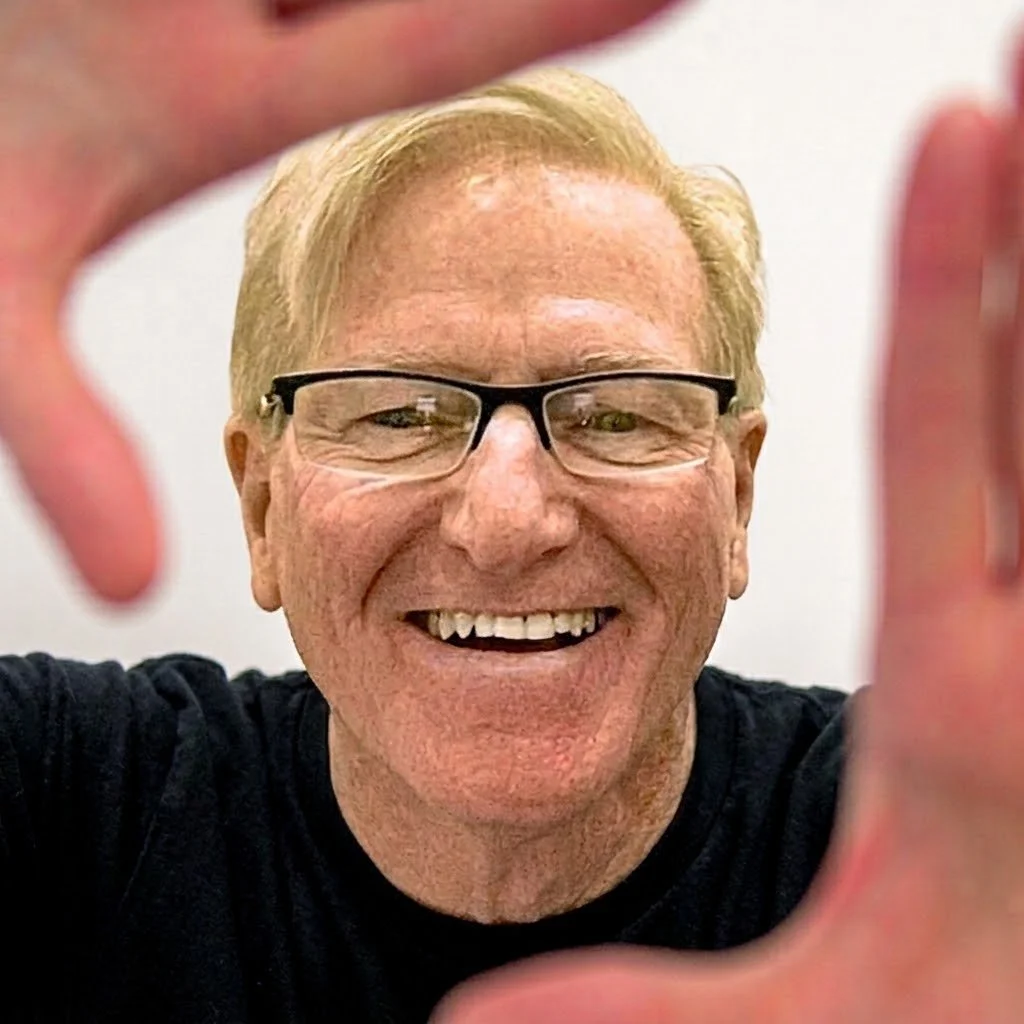 A smiling man with glasses and blonde hair, holding the camera with his hands framing his face in front of a plain background.