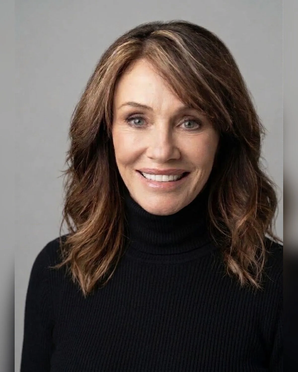 A woman with shoulder-length wavy brown hair wearing a black turtleneck sweater, smiling against a gray background.