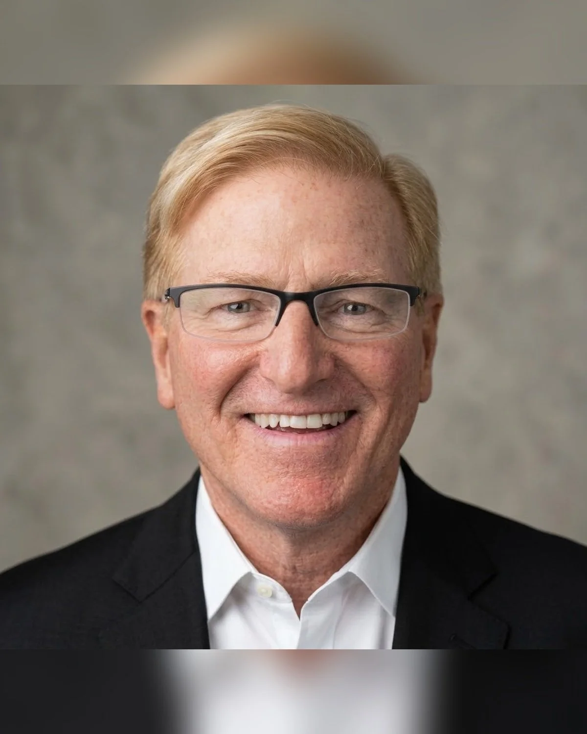 Professional headshot of a smiling man with glasses, light-colored hair, wearing a black suit and white shirt, against a gray background.