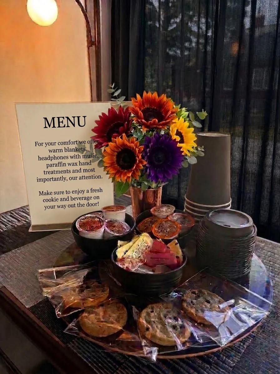 Table with a menu sign, colorful sunflower bouquet, candles, stacked black paper plates, and assorted snacks including cookies and candies.
