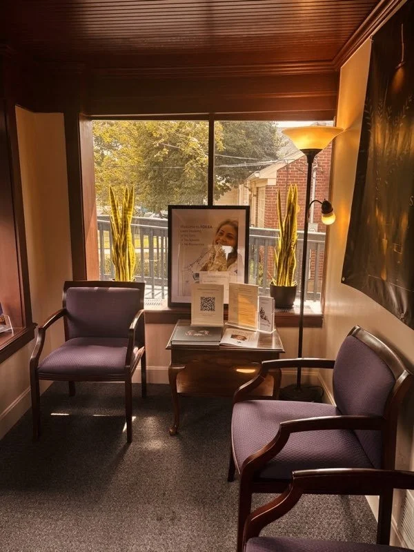 A cozy waiting area with two purple upholstered chairs, a small wooden table displaying brochures and flyers, decorative yellow plants, a standing lamp, and a large window letting in natural light.