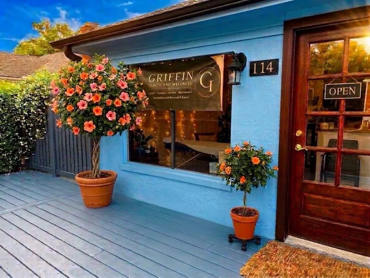 The exterior of a health and wellness business called Griffin Health and Wellness, with a large window, a wooden door with a glass panel and a sign that says "OPEN," and two potted flowering plants with orange blossoms flanking the entrance. The building is painted light blue, and there is a black sign above the window with the business name and contact details, and the house number 114."}