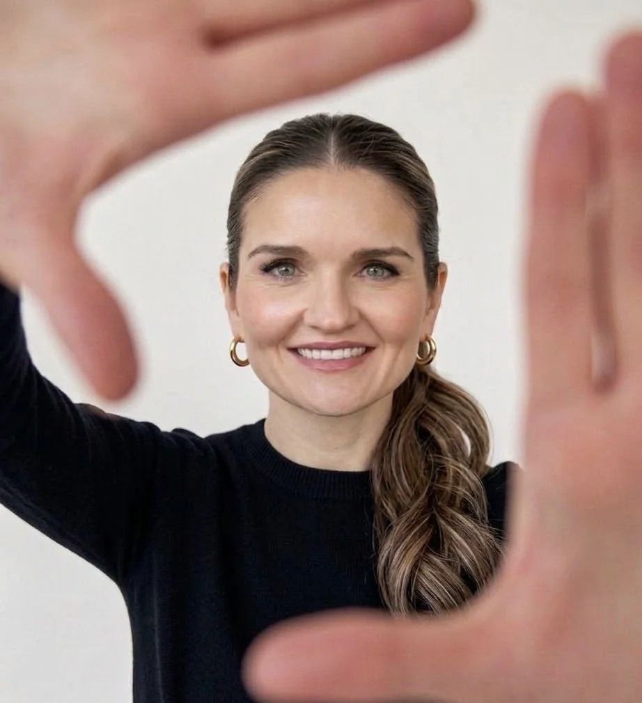 A woman with long brown hair, wearing a black top and gold hoop earrings, smiling and framing her face with her hands in a rectangle shape.
