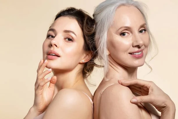 Two women with light skin and different hair colors posing together, one with dark hair and the other with gray hair, against a plain beige background.