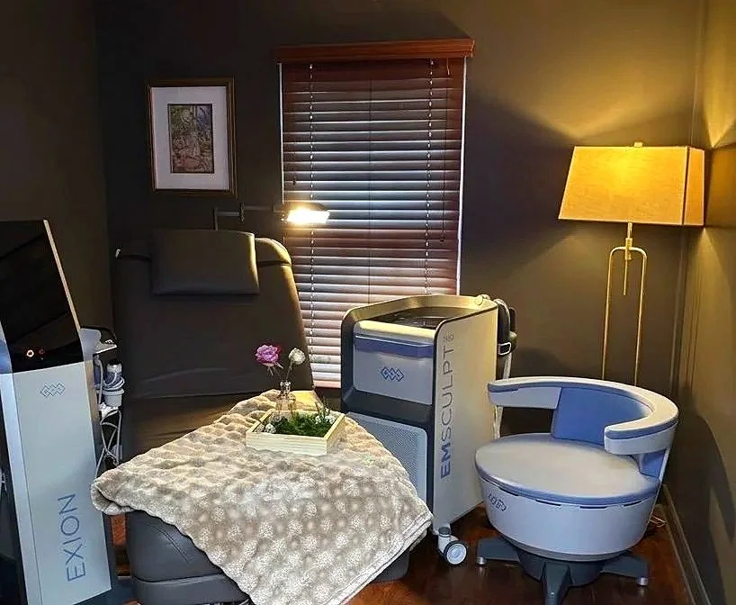 A spa or wellness room featuring massage or therapy chairs, a side table with a flower arrangement, and a medical or aesthetic treatment device. The room has a window with blinds, a framed picture on the wall, and lamps providing warm lighting.