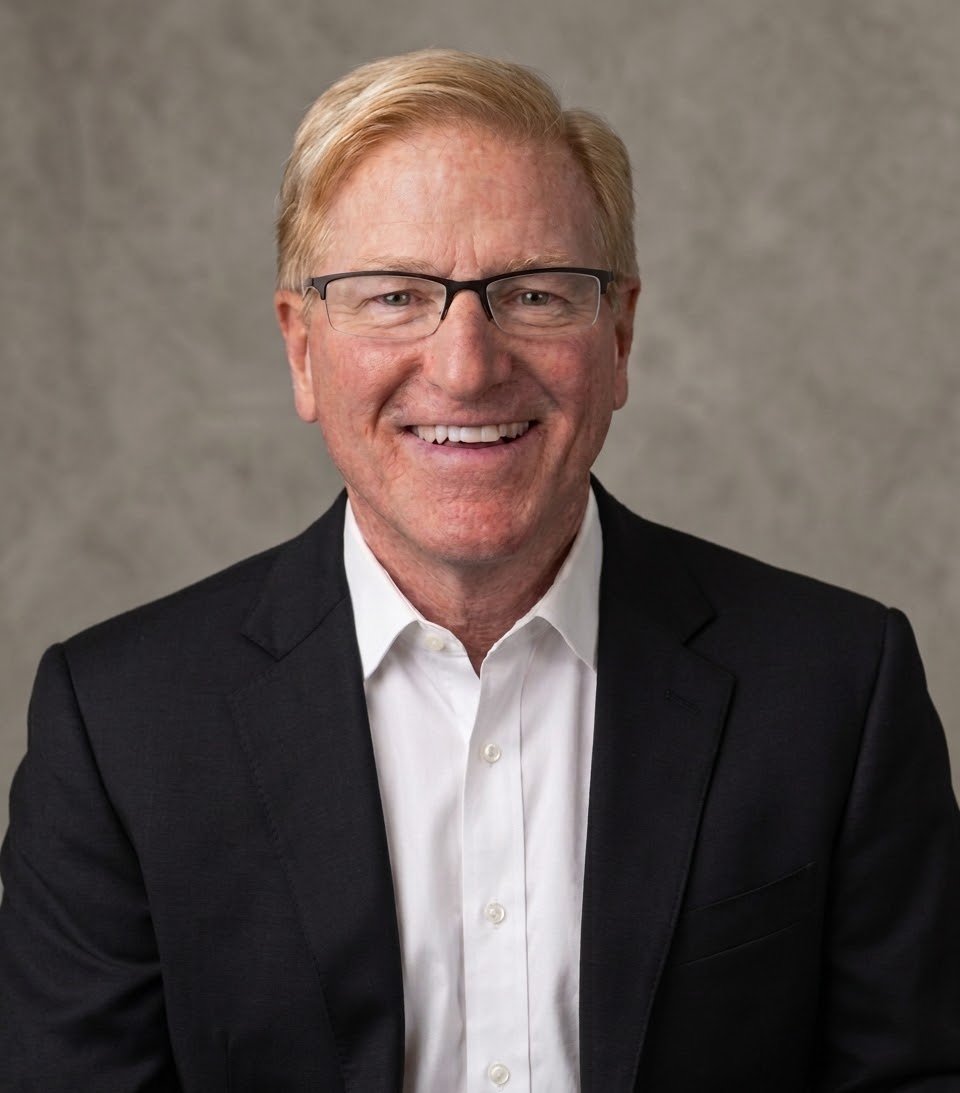 A smiling middle-aged man with blonde hair, wearing glasses, a white dress shirt, and a black blazer, posed against a neutral gray background.