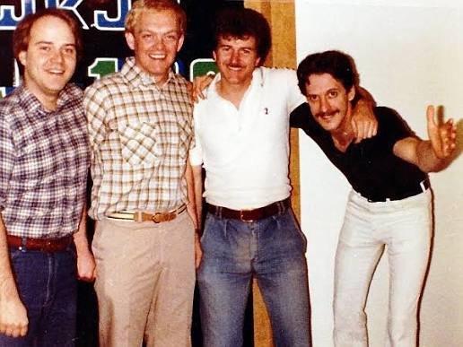 Four men standing together in an indoor setting, smiling at the camera, with the man on the far right leaning forward and making a peace sign.