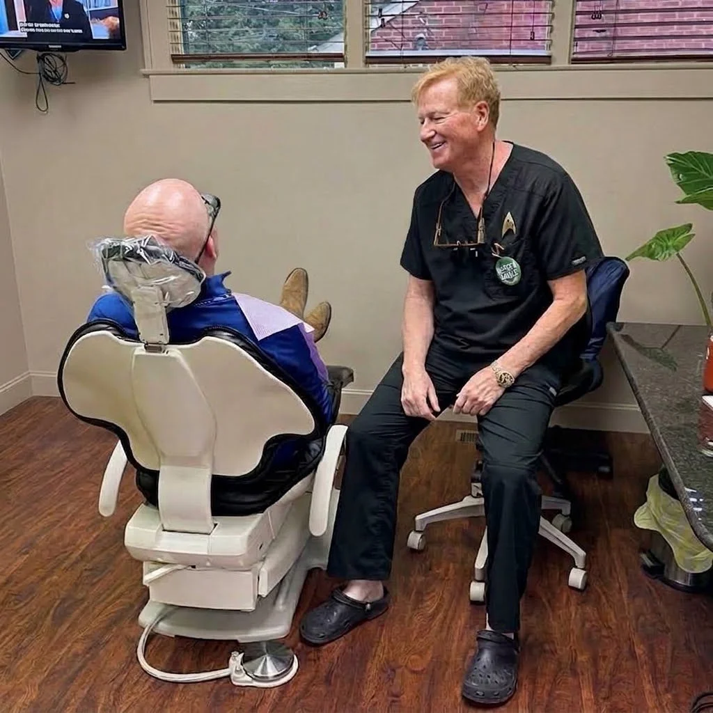 A man and woman in a dentist's office, smiling and engaging in conversation. The woman is seated in a dental chair, and the man is sitting on an office chair beside her, both appearing happy and relaxed.